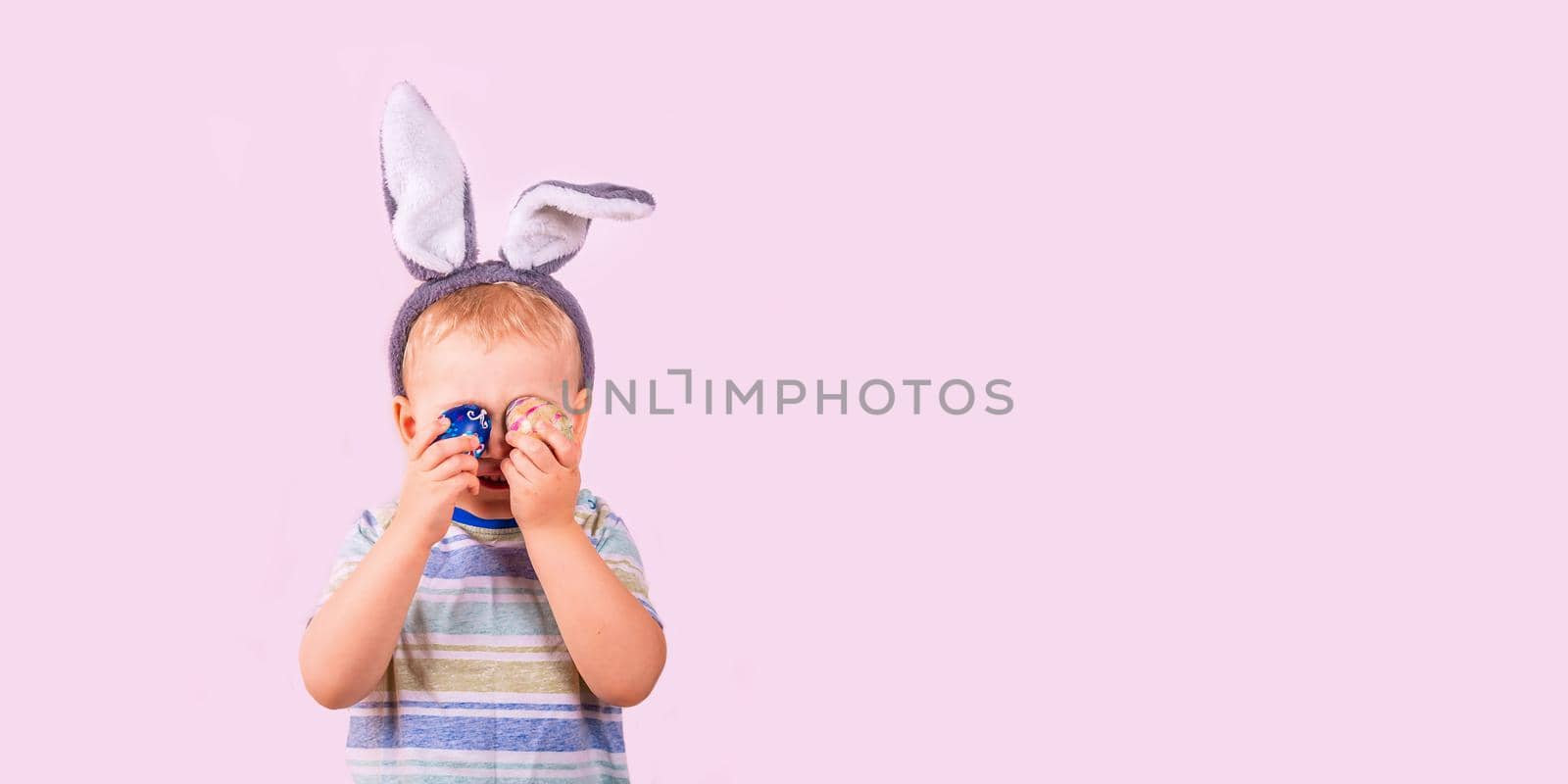 Cute baby boy in rabbit bunny ears on head closing his eyes with colored eggs on pink background. Easter holiday by Len44ik