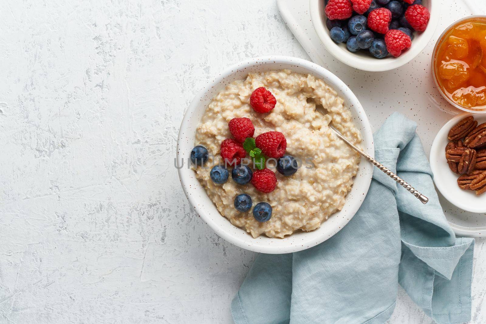 Oatmeal rustic porridge with blueberry, raspberries, jam and nuts in white bowl, dash diet with berries, white background, top view copy space. Healthy diet breakfast