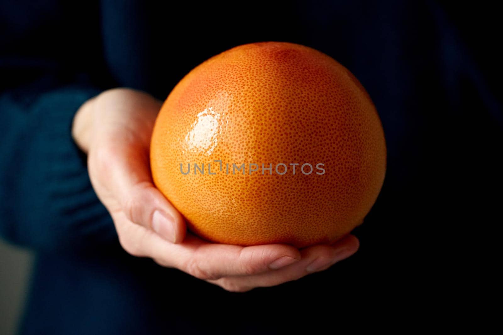 Close-up of woman's hand holding whole bright citrus fruit grapefruit on dark background. Lifestyle, natural light from window. Vitamins