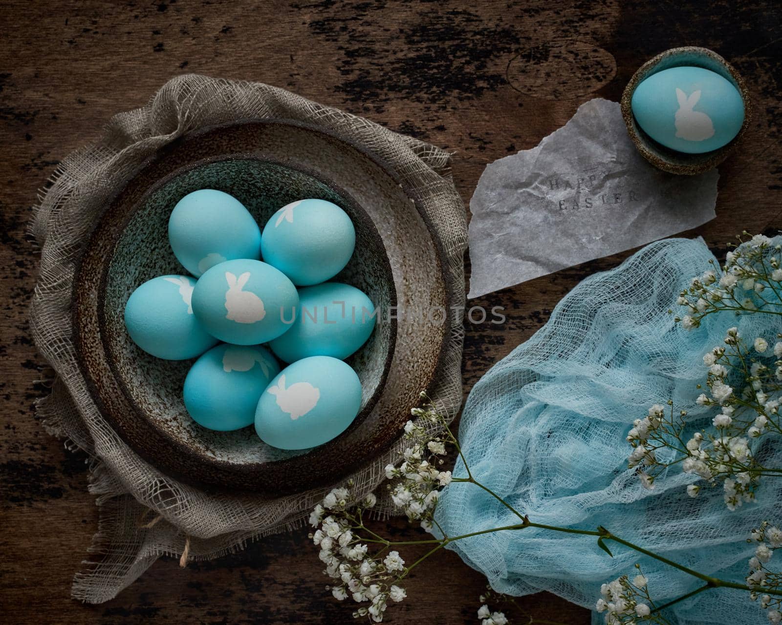 Unusual Easter on dark old background. Ceramic brown bowl with blue eggs with rabbit. Darkness, rays of sunlight shine on eggs. Concept of new life, rebirth. Rustic style. Top view