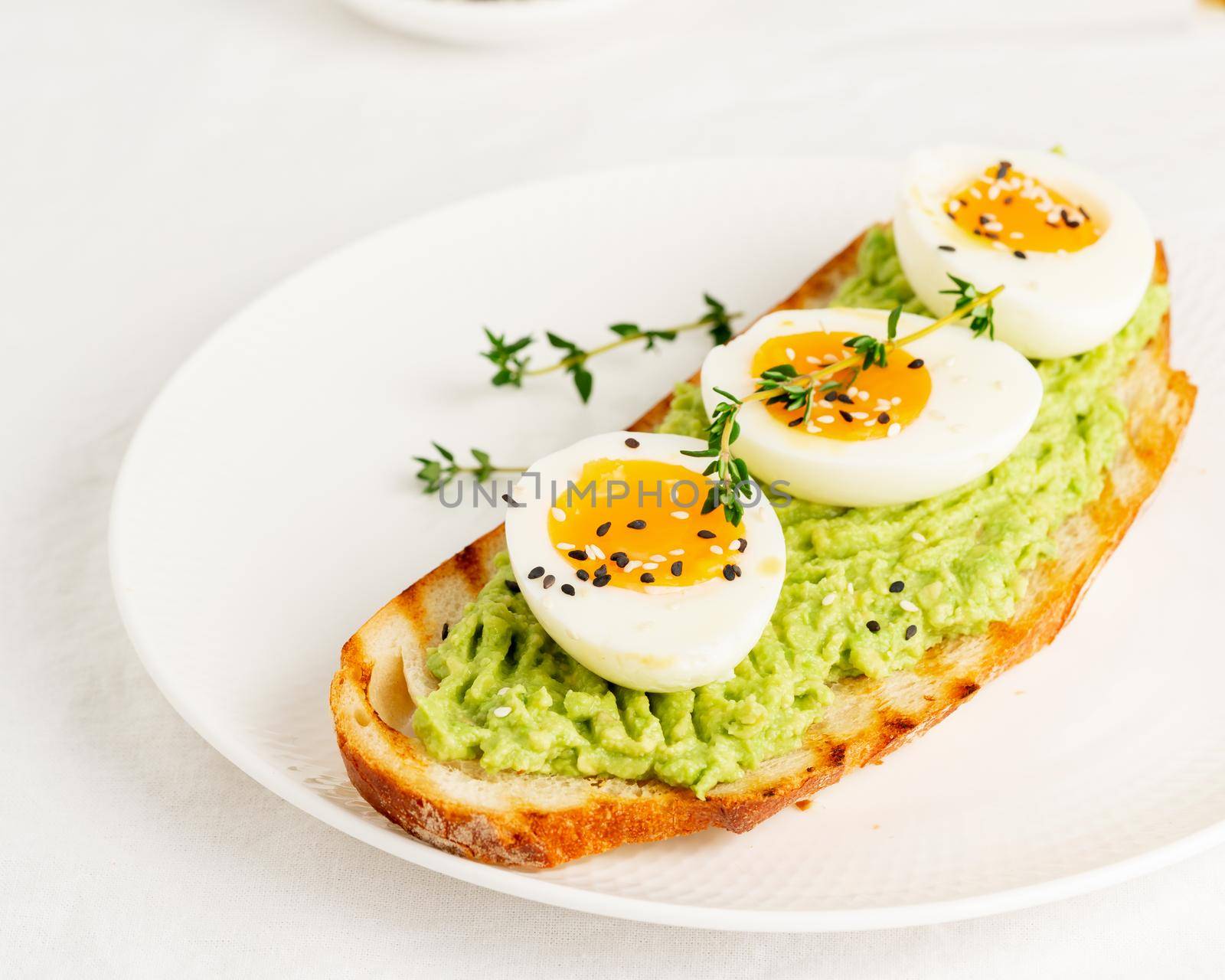 Avocado toast with toasted bread soft-boiled eggs with yellow yolk and tomatoes with herbs on white plate on light tablecloth