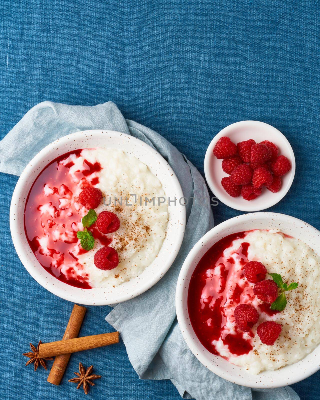 Rice pudding. French milk rice dessert with raspberries, blueberries, berries, jam. Healthy Vegan diet breakfast with coconut milk, cinnamon. Blue linen textile. Top view, vertical, copy space