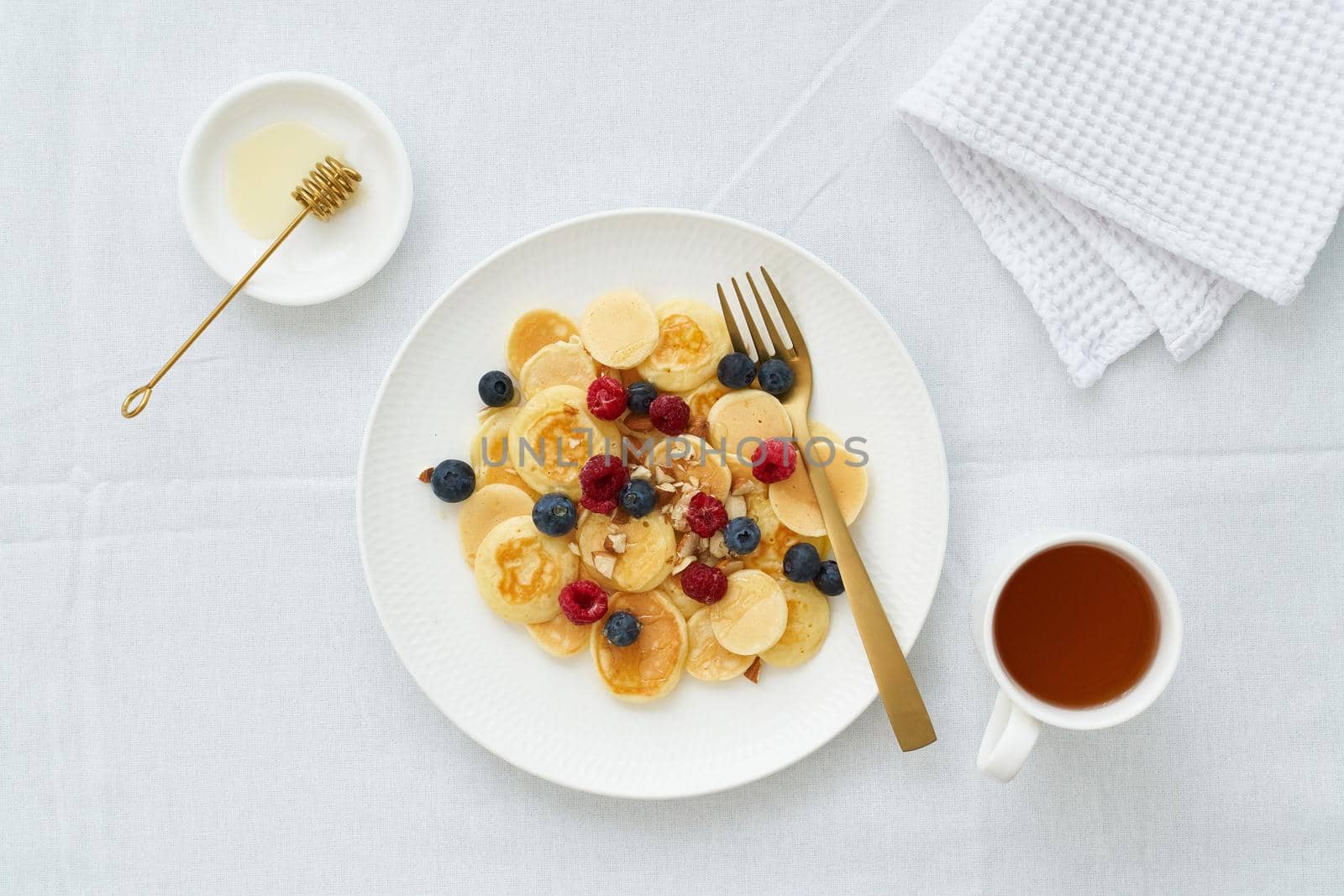 Tiny pancakes, small thin funny crumpet, children's food. Breakfast with raspberries, blueberries, honey and nuts, white tablecloth, bright morning, top view