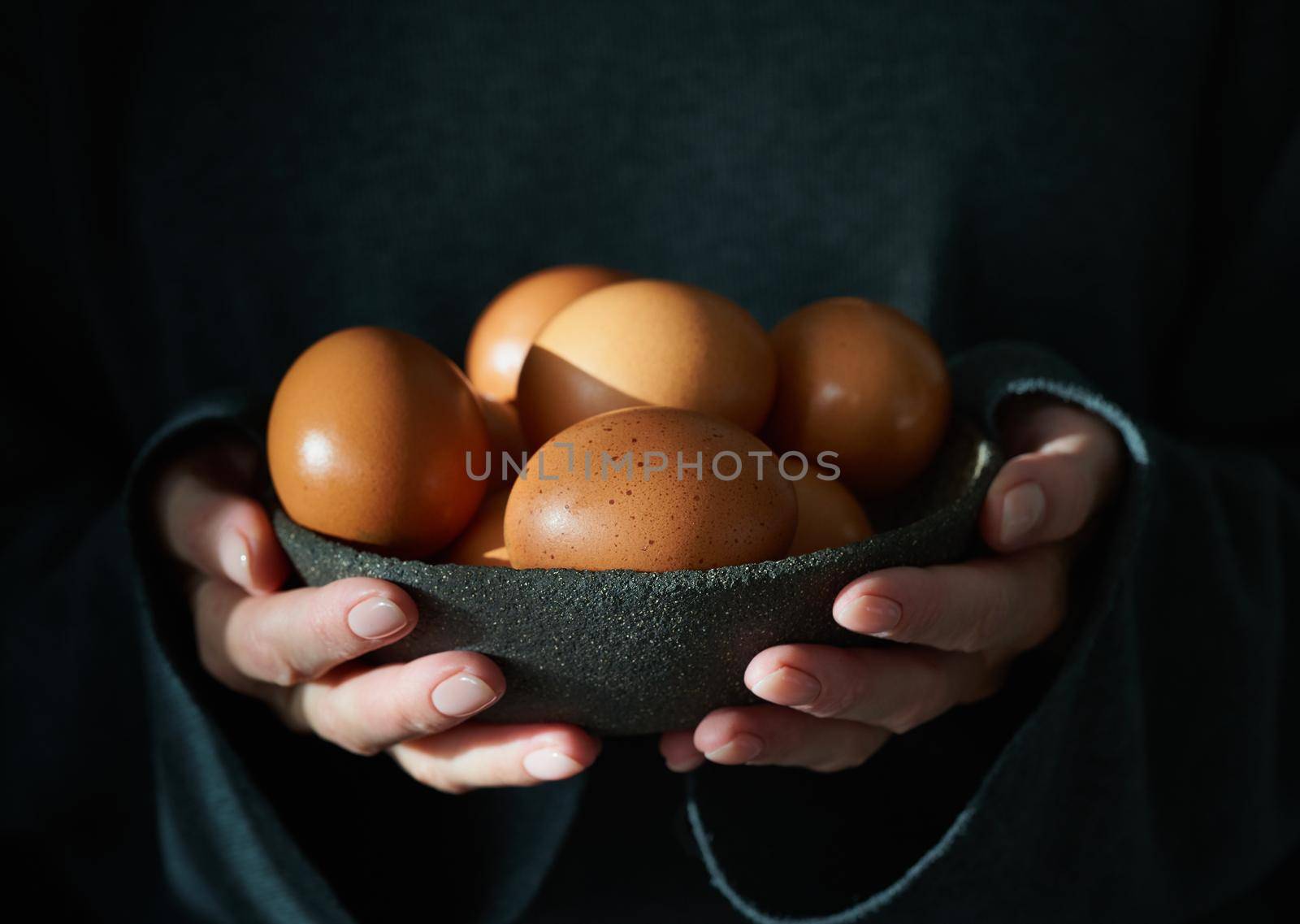 Unusual Easter on dark background. Bowl of brown eggs with hands. Darkness, rays of sunlight shine on the eggs. Concept of new life, rebirth. Rustic style. Side view, copy space