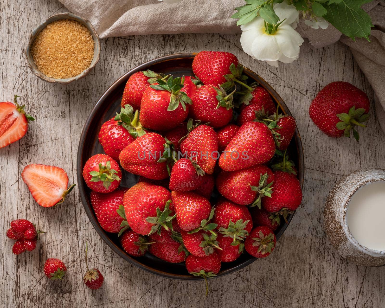 Top view of bowl with strawberry on wooden table, bottle with milk, flower. Dark still-life photo with summer berries, vertical