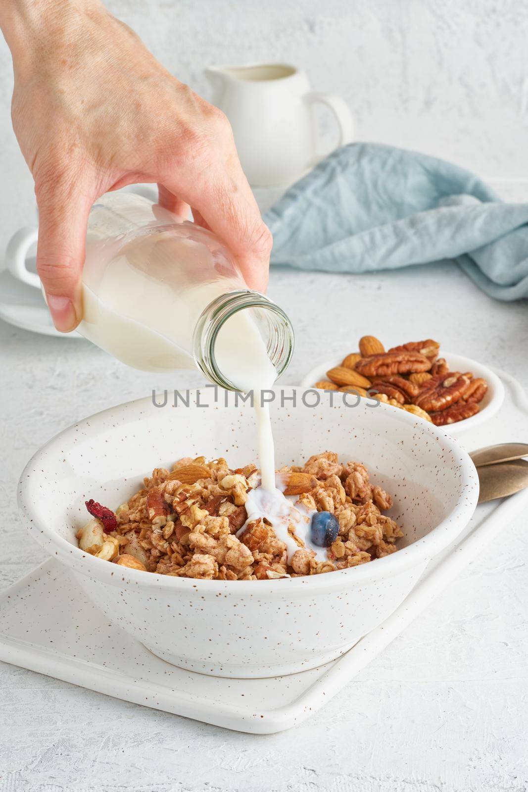 Granola. Breakfast, healthy diet food with oat flakes, nuts, banana, white table. Woman's hand pours milk into muesli from bottle, vertical