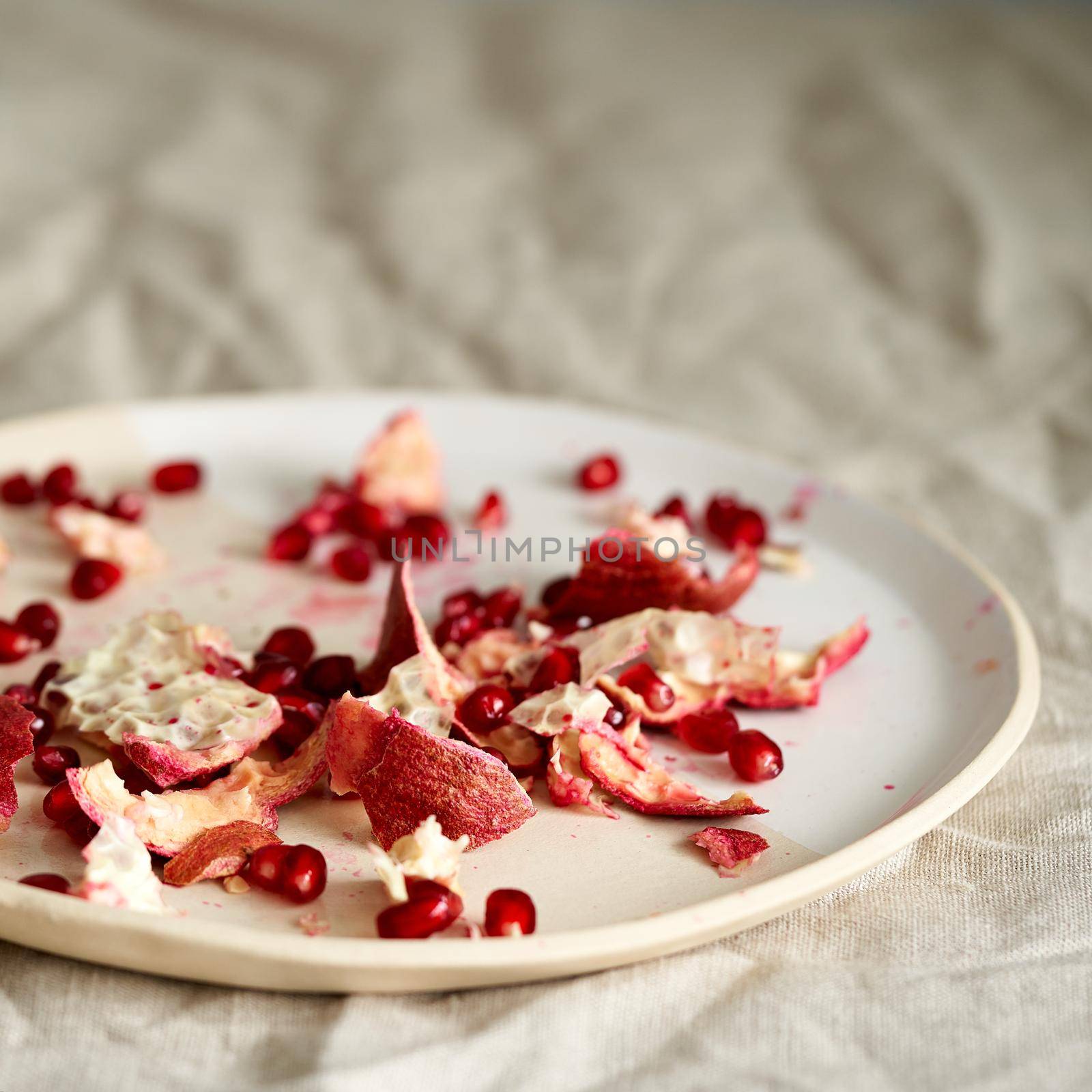 food waste of pomegranate cleaning and seeds on plate on table covered with crumpled beige tablecloth in natural light from window. Lifestyle photo