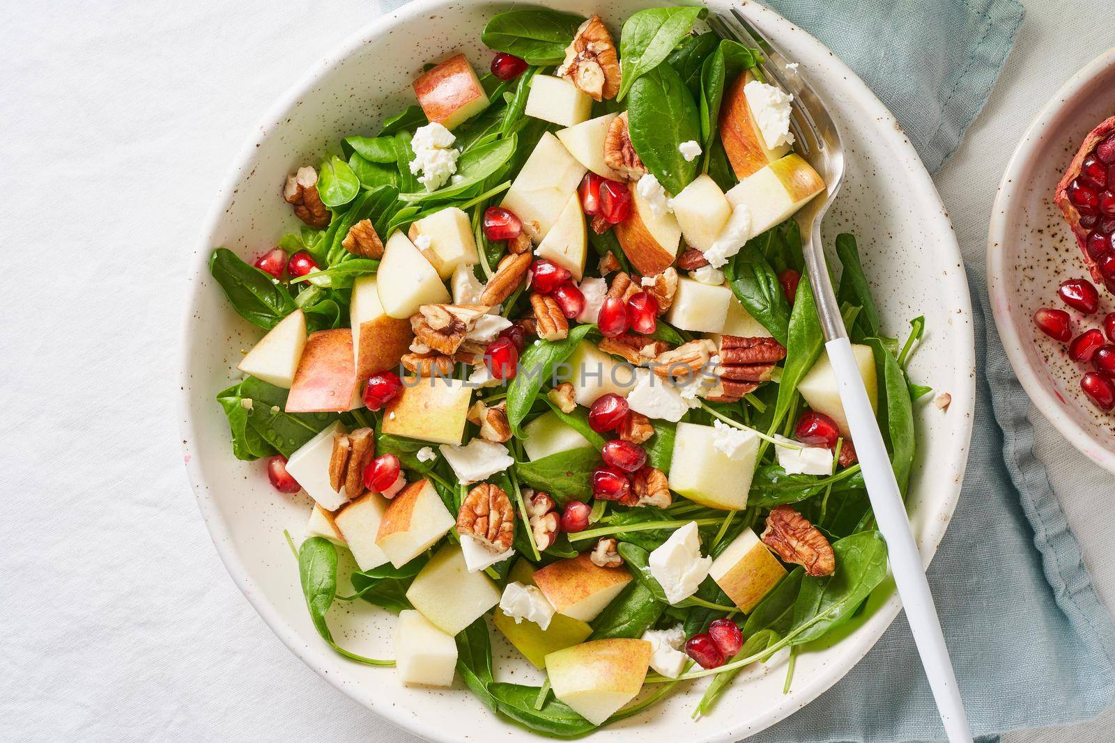 Fruits salad with nuts, balanced food, clean eating. Spinach with apples, pecans and feta, garnished with pomegranate seeds in bowl on table with white tablecloth.