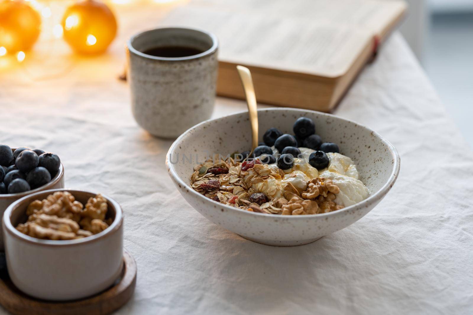 Reading book and eating healthy Christmas holiday winter breakfast with granola muesli and yogurt in bowl on white table background, lights garlands in bokeh. Organic morning diet meal with oat