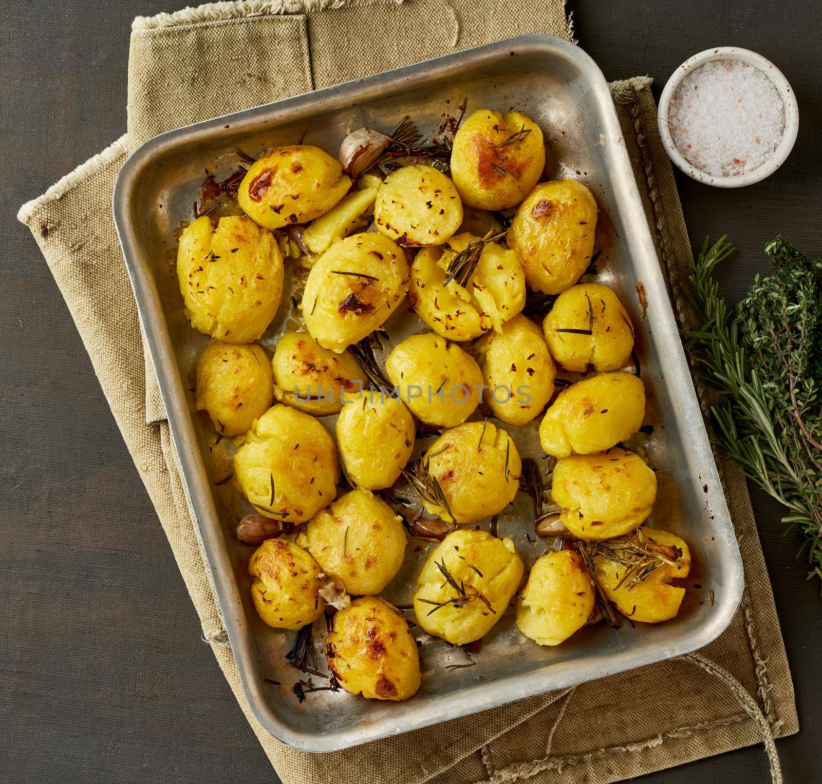 Oven baked whole crushed and crusty potato spuds with seasoning and herbs in metalic tray. Roasted smashed potatoes. Top view. Dark rustic table.