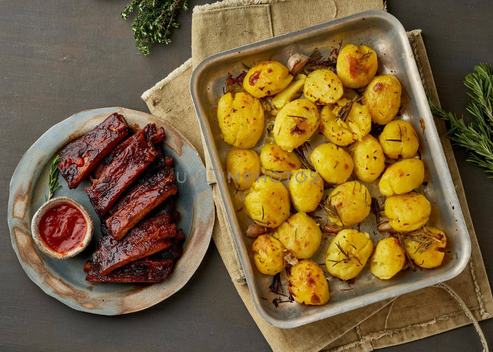 Barbecue pork ribs and crushed smashed potatoes. Slow cooking recipe. Whole pickled roasted pork meat with red sauce, side view, close up. Dark background