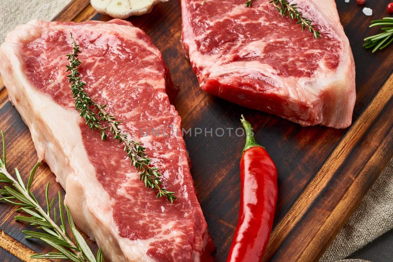 Two big whole piece of raw beef meat, striploin on wooden cutting board on dark brown background, macro, close up. Seasoning steak with salt, thyme, garlic