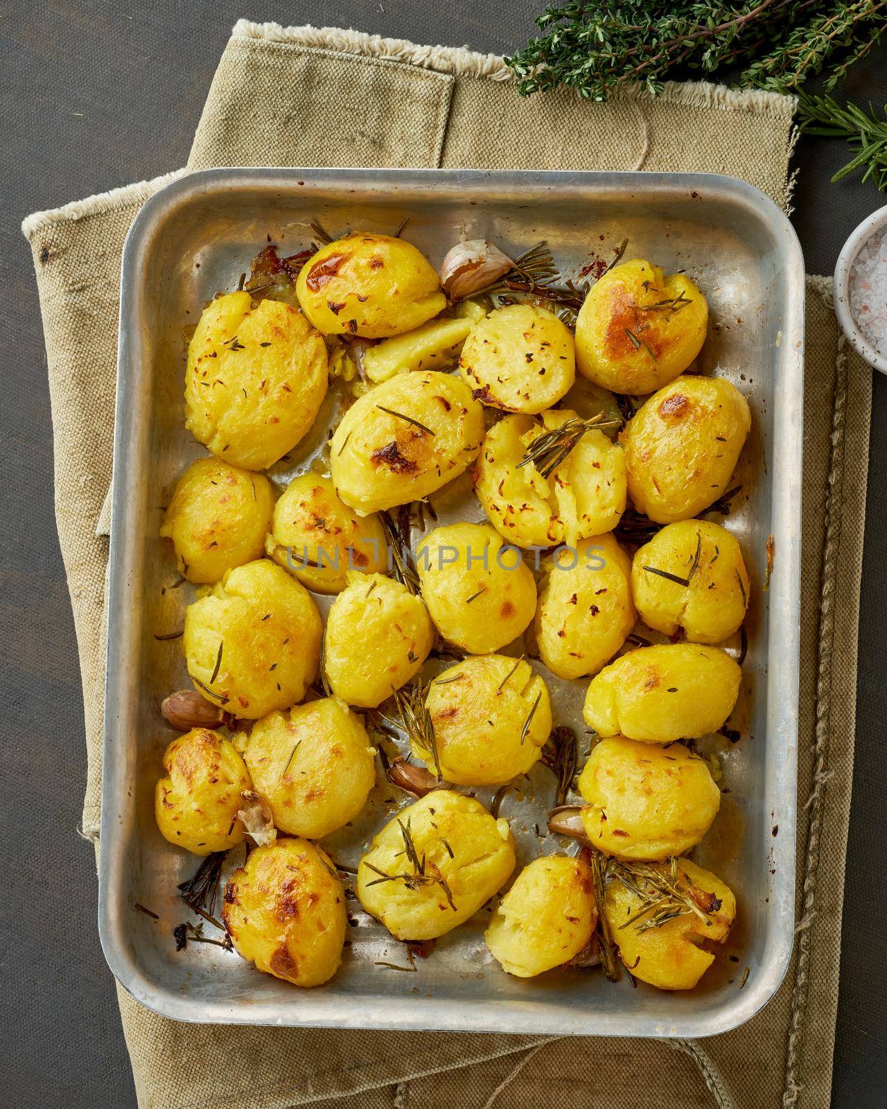 Oven baked whole crushed and crusty potato spuds with seasoning and herbs in metalic tray. Roasted smashed potatoes. Top view. Dark rustic table. Vertical