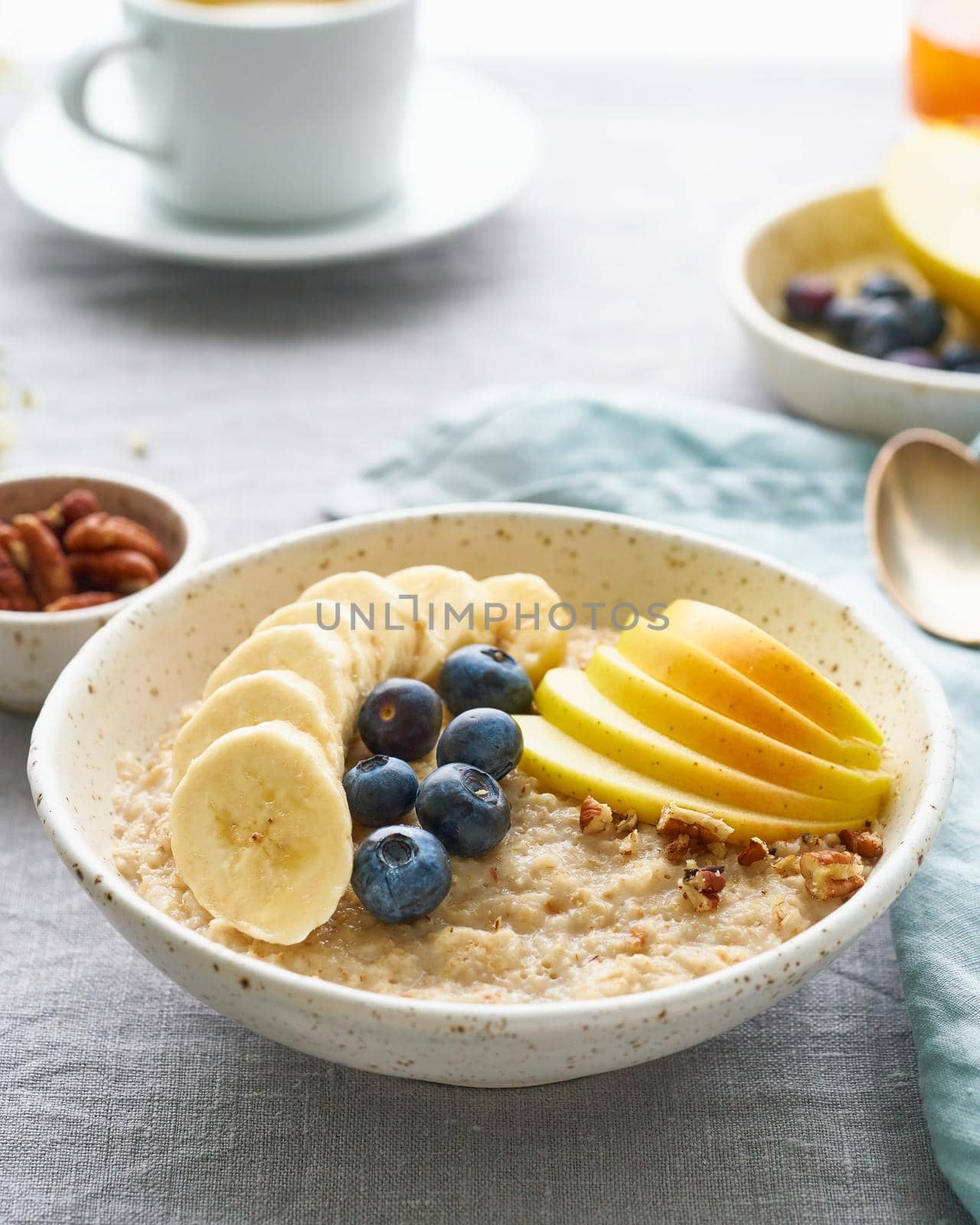 Whole oatmeal, large bowl of porridge with banana, blueberries, apple, nuts for breakfast, morning meal. Side view, close up, vertical, gray table. Vegan tasty healthy diet.