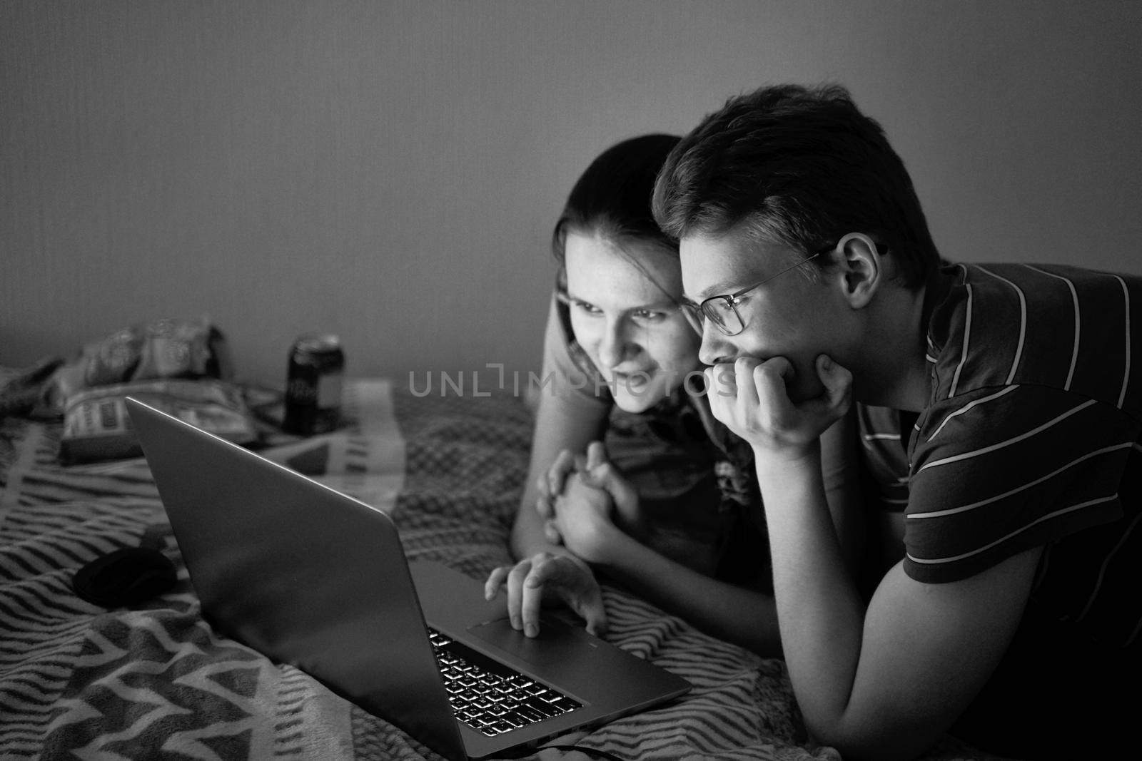 Two teenagers watching film during quarantine due to coronavirus pandemic. Boy and girl look at laptop while staying at home. Dark, evening