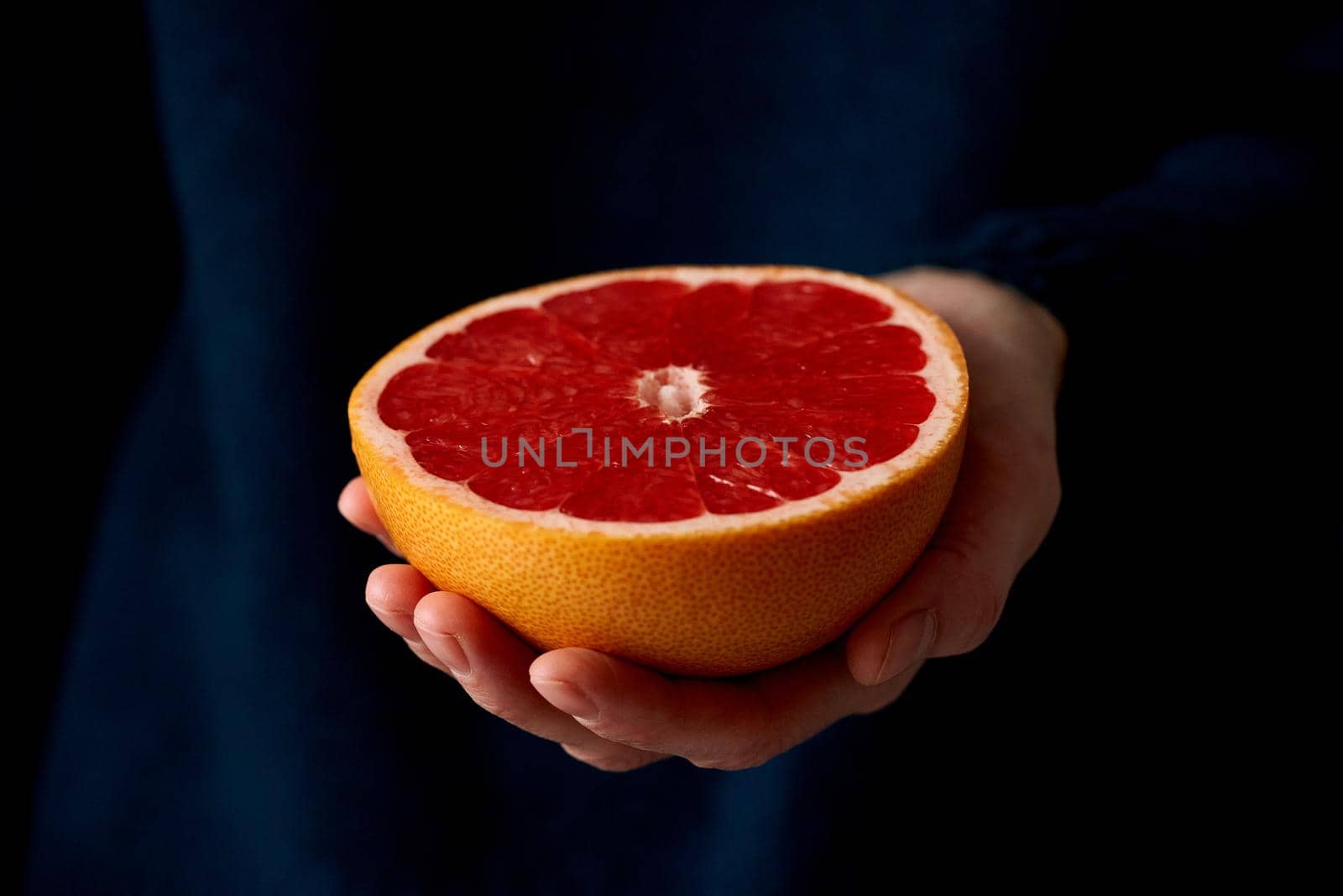 Close-up of woman's hand holding fresh halved red bright citrus fruit grapefruit on dark background. Lifestyle, natural light from window. Vitamins
