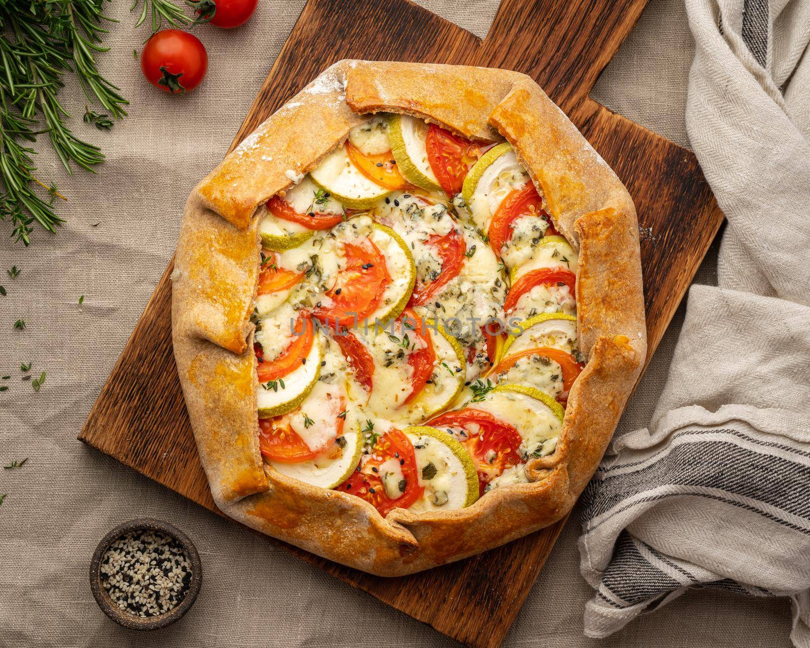 Homemade savory galette with vegetables, wheat pie with tomatoes, zucchini, blue cheese Gorgonzola. Rustic crust crostata on dark linen textile tablecloth. Top view