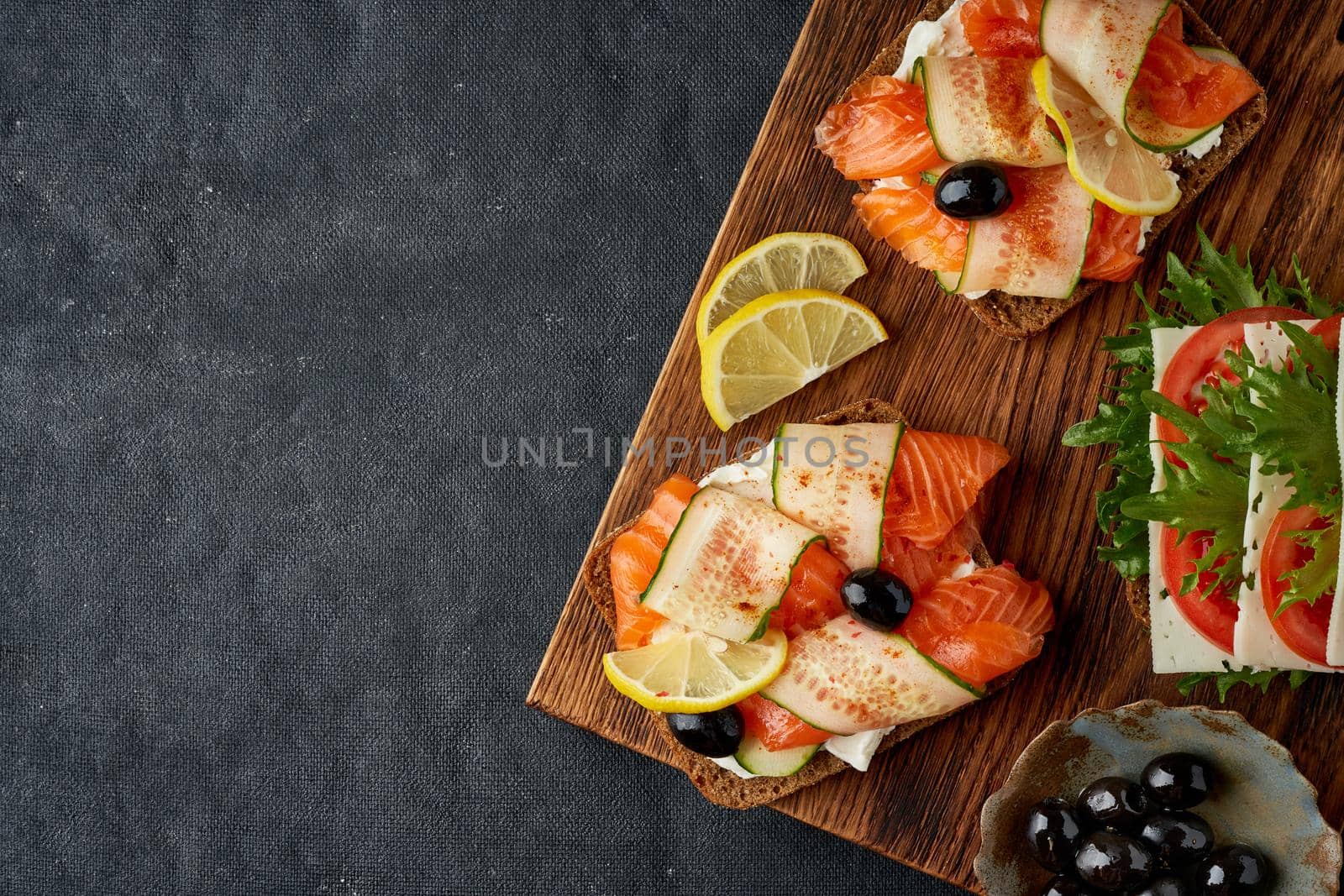 Smorrebrod - traditional Danish sandwiches. Black rye bread with salmon, cream cheese, cucumber, tomatoes on dark brown wooden background, copy space