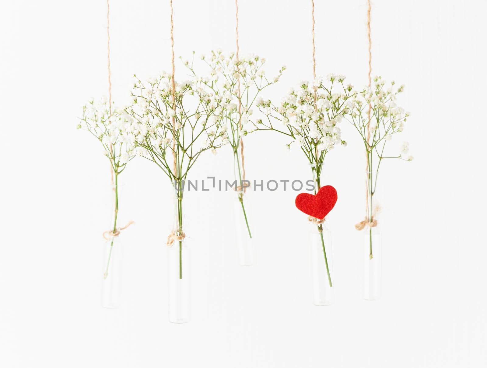 White flowers in glass mini vases hanging on white background. Red heart is symbol of love. Concept of Valentine's Day, wedding celebration. Copy space