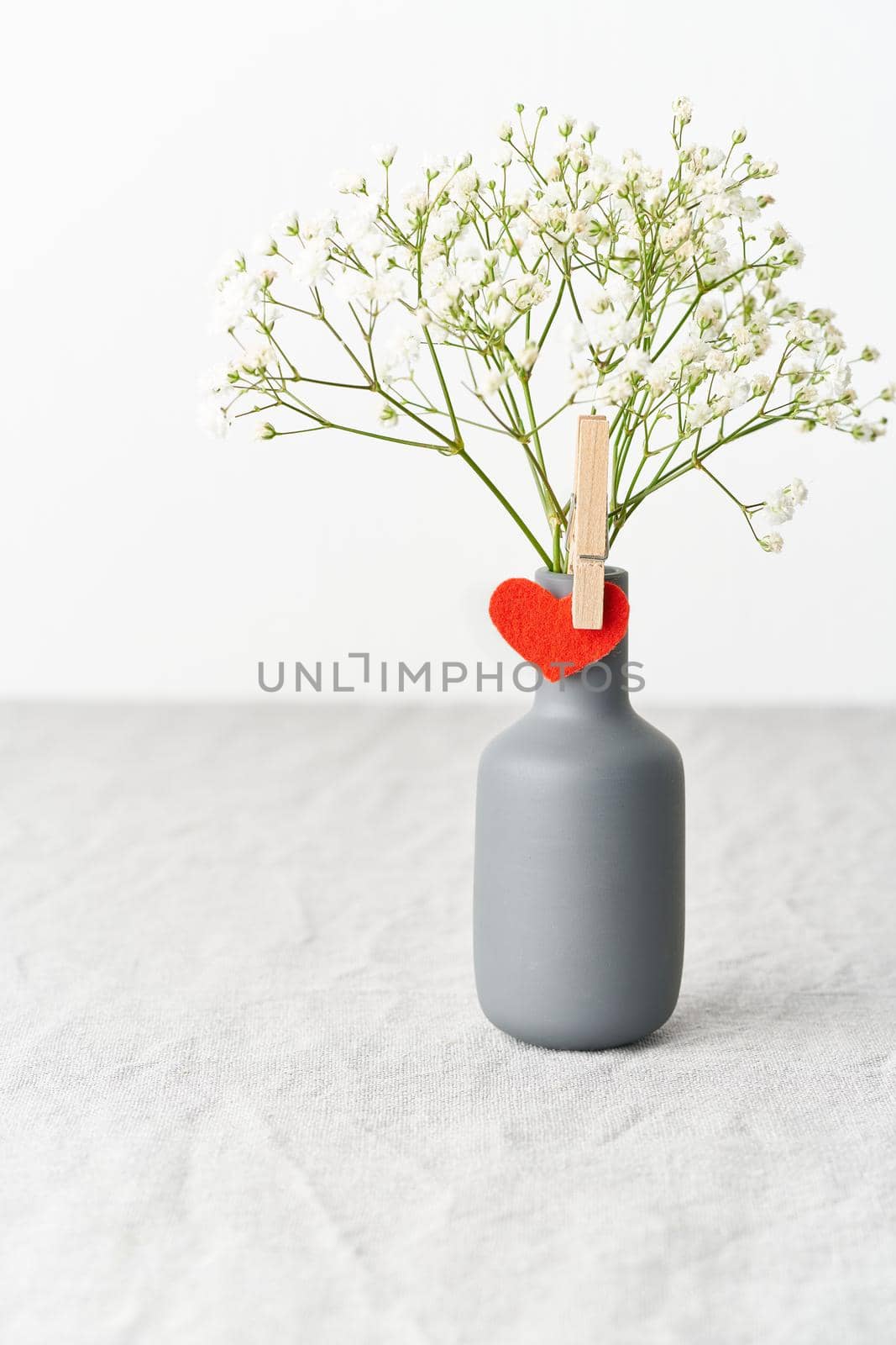 Valentine's Day. Delicate white flowers in a vase. Red felt heart - symbol of lovers. Light white gentle pastel background. Scandinavian minimalism. Side view, copy space, vertical