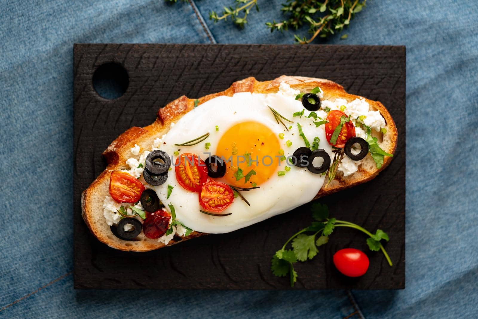 Toasted bread toast with fried eggs with yellow yolk and tomatoes, olives, sprinkled with herbs on dark wooden serving board on blue denim napkin, top view