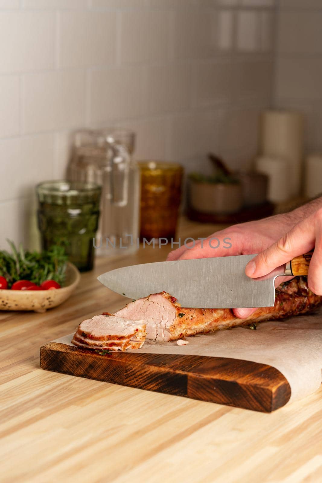 Unrecognizable man cuting with large knife baked pork tenderloin on cutting board on itchen table at home, slicing thin slices of meat with sharp blade, vertical