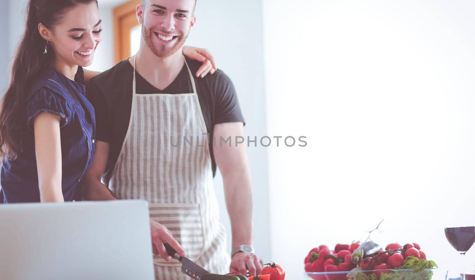 Young man cutting vegetables and woman standing with laptop in the kitchen.
