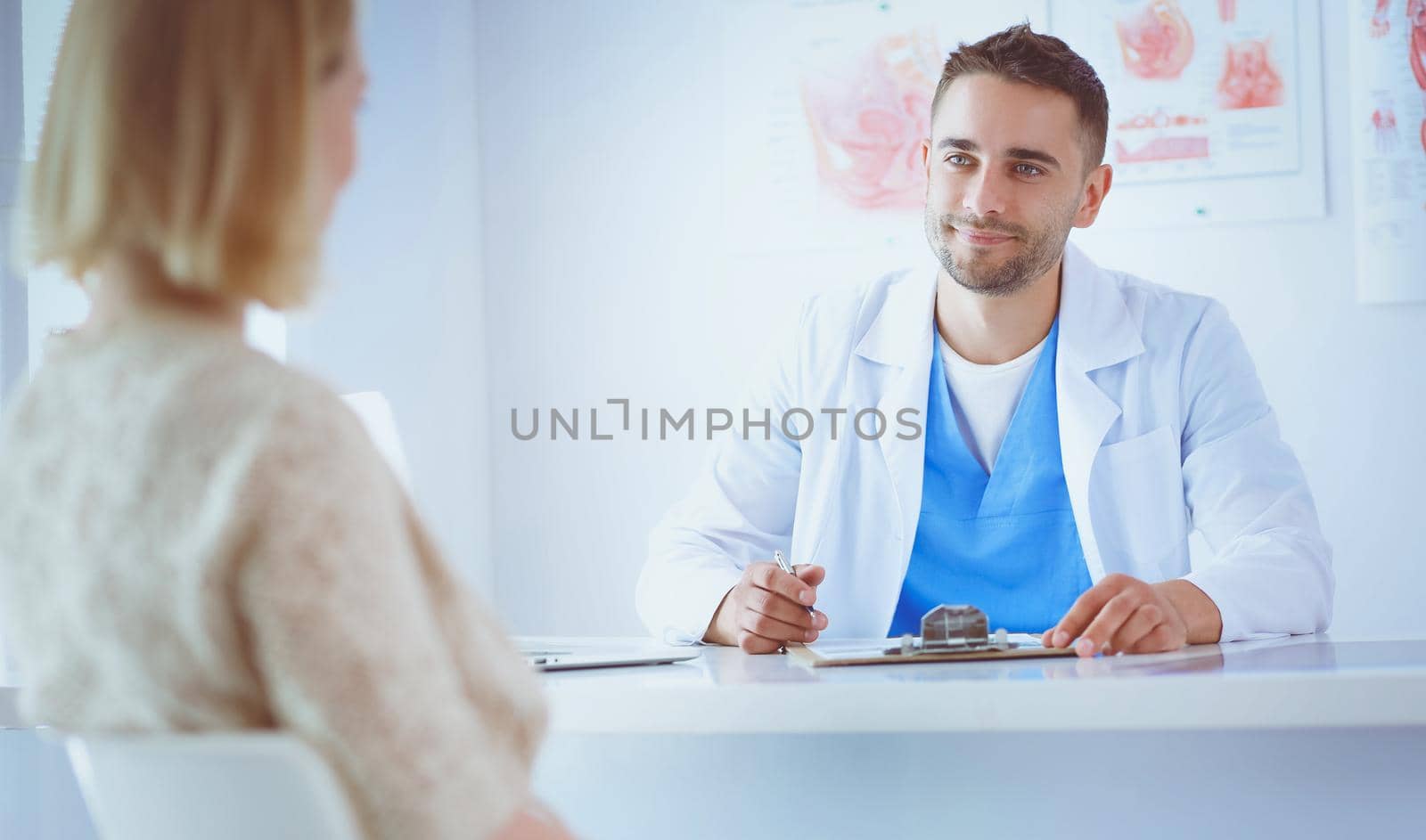 Handsome doctor is talking with young female patient and making notes while sitting in his office.