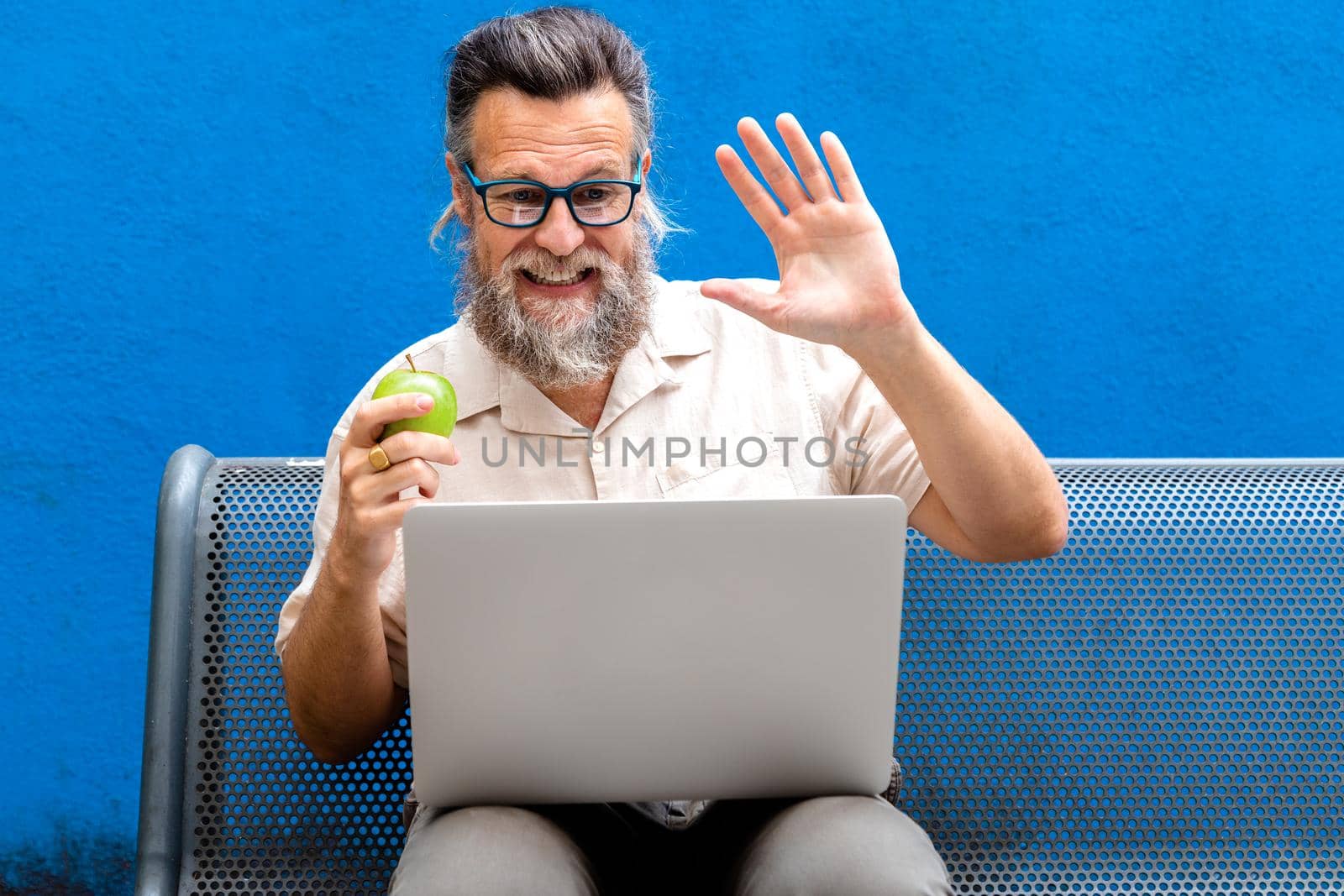 Mature caucasian man eating apple waving hello on a video call in park bench. Lifestyle and technology concepts.