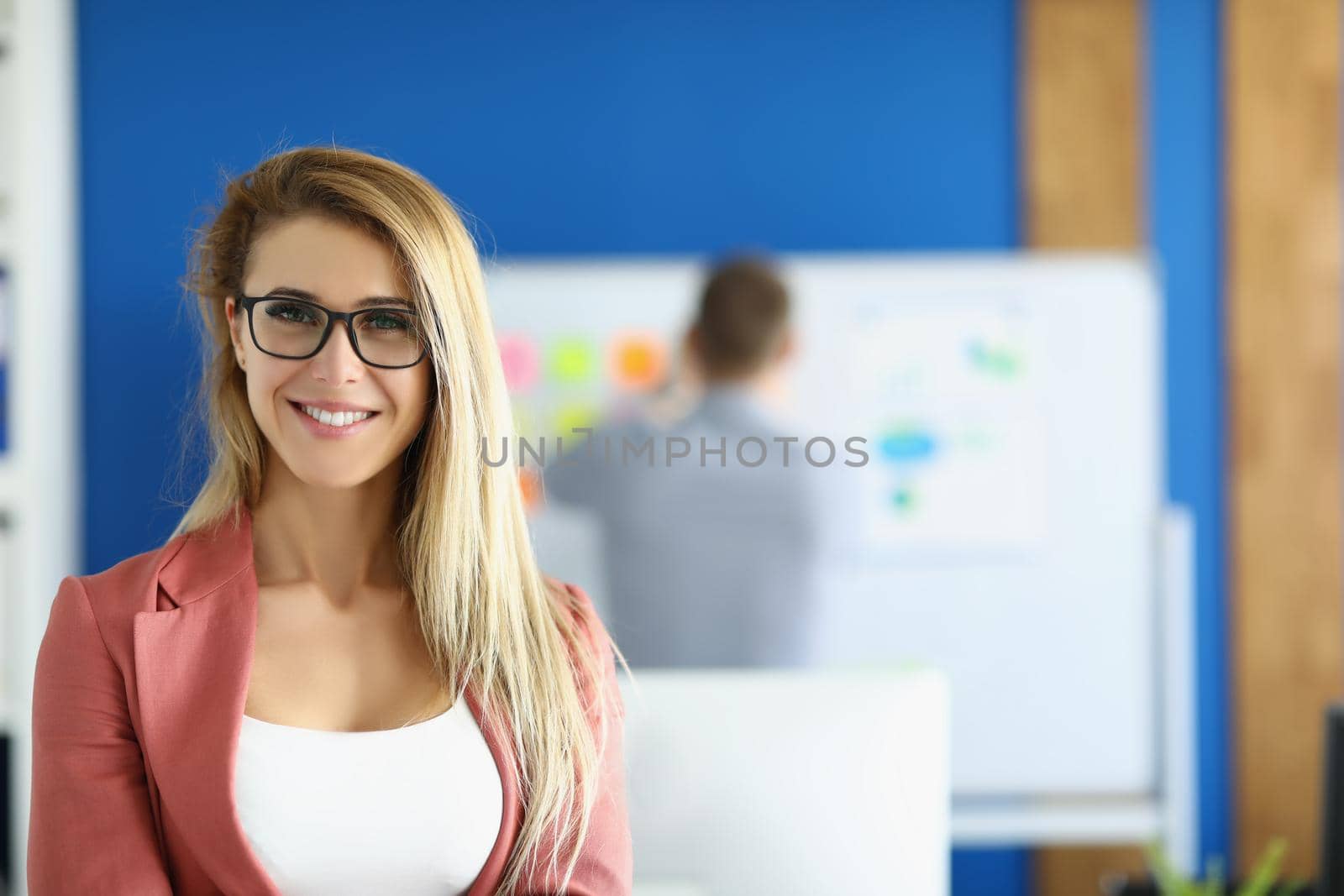Portrait of smiling businesswoman posing in office with in presentable pink costume, colleague on background. Business, office life, career growth concept