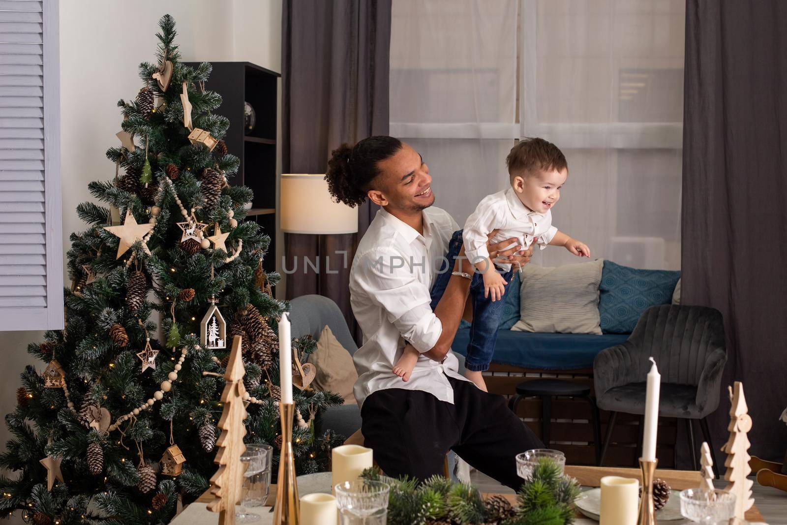 A young father is holding his little son in outstretched arms, playing in the living room. There is a Christmas tree with wooden toys nearby