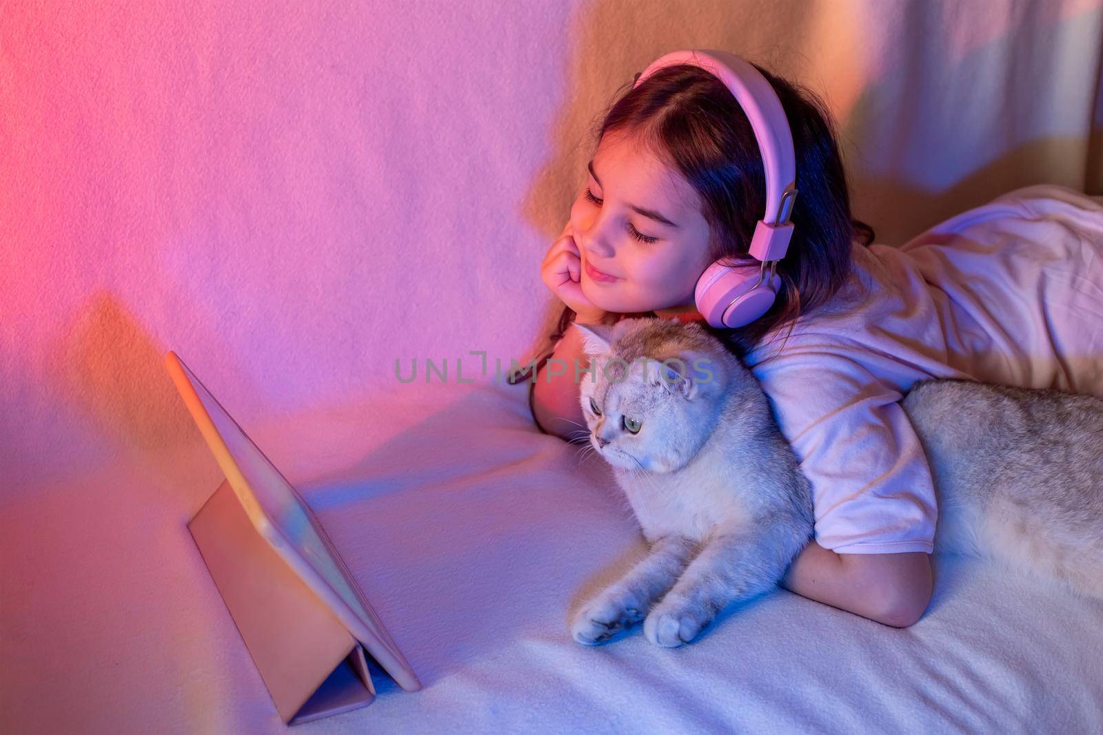 A happy little girl in pink headphones, lies on the bed, with a white adorable cat, in a neon pink-blue light, looks into a digital tablet