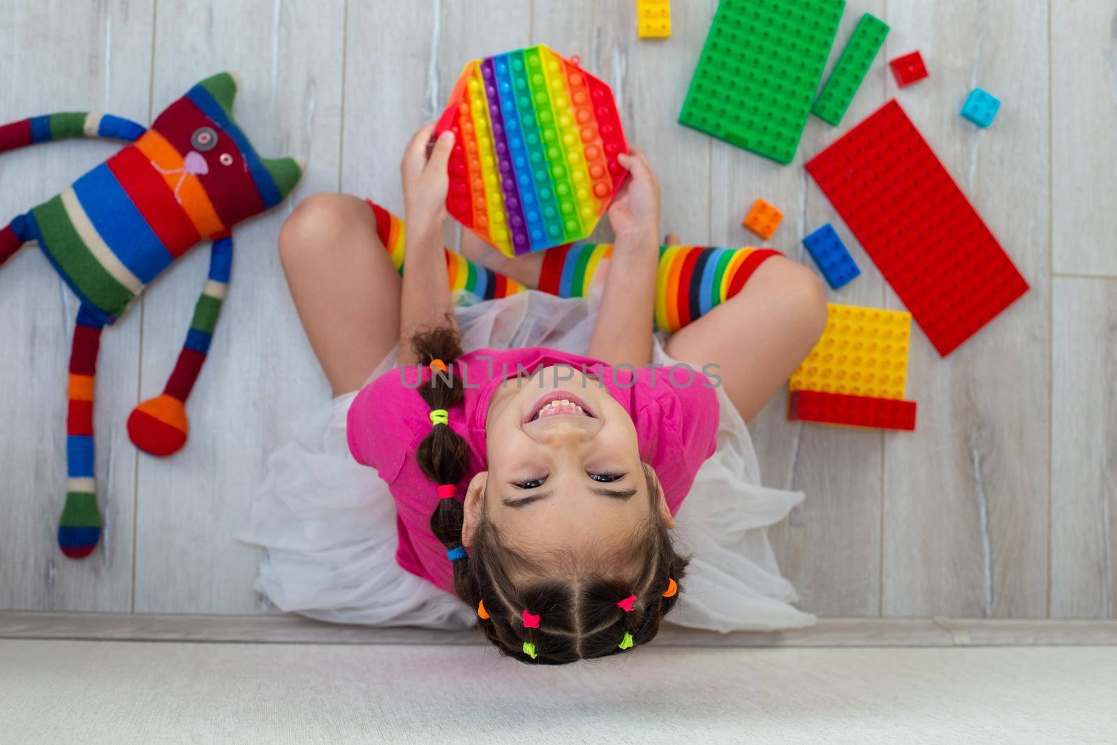 A cheerful little brunette in a pink T-shirt and rainbow long socks sits on the floor surrounded by scattered colorful toys and holds a rainbow antistress toy in her hands. Looks up into the camera.