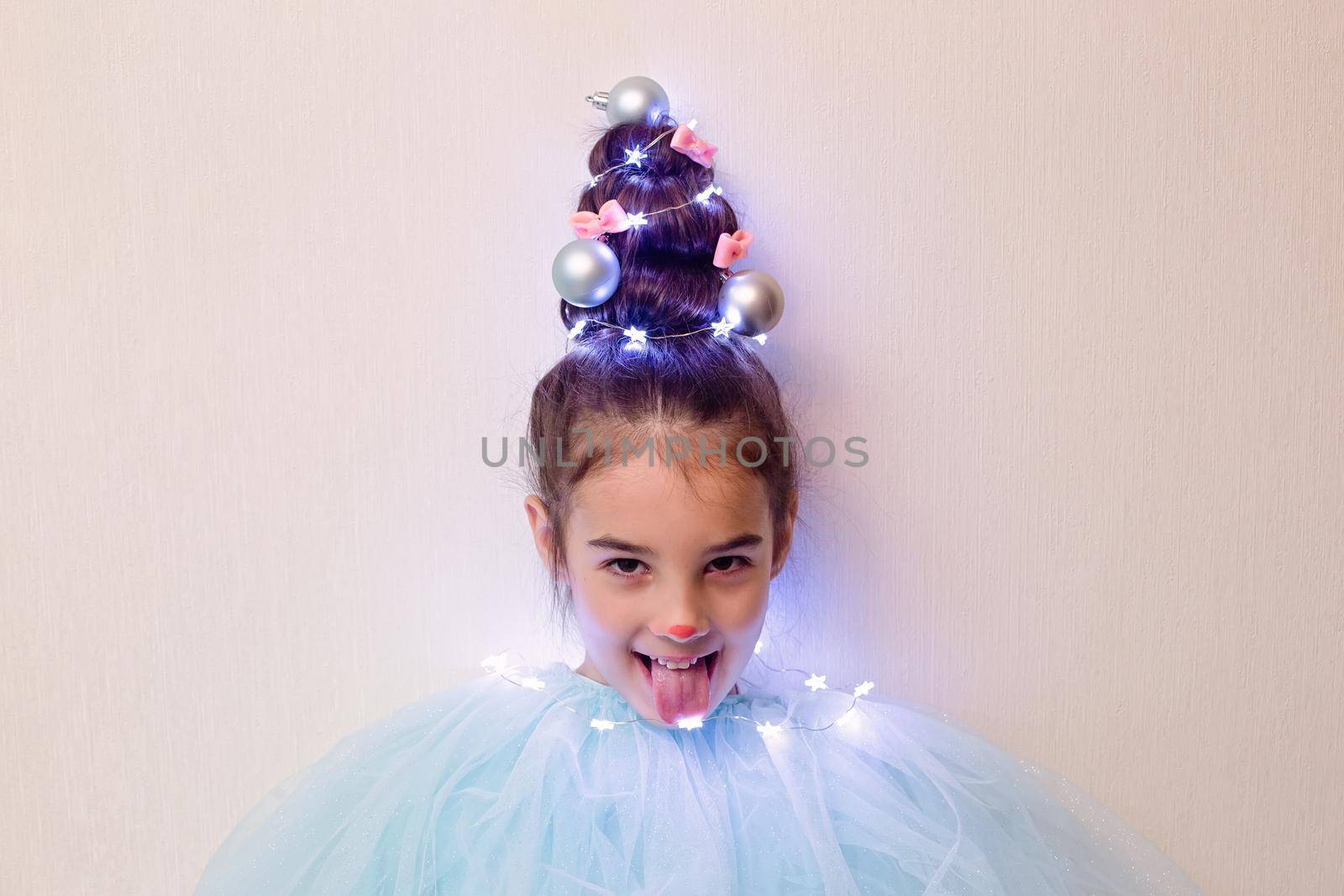 A funny brunette girl with a hairstyle in the form of a Christmas tree with a luminous garland, stands against the background of a light wall, sticking out her tongue