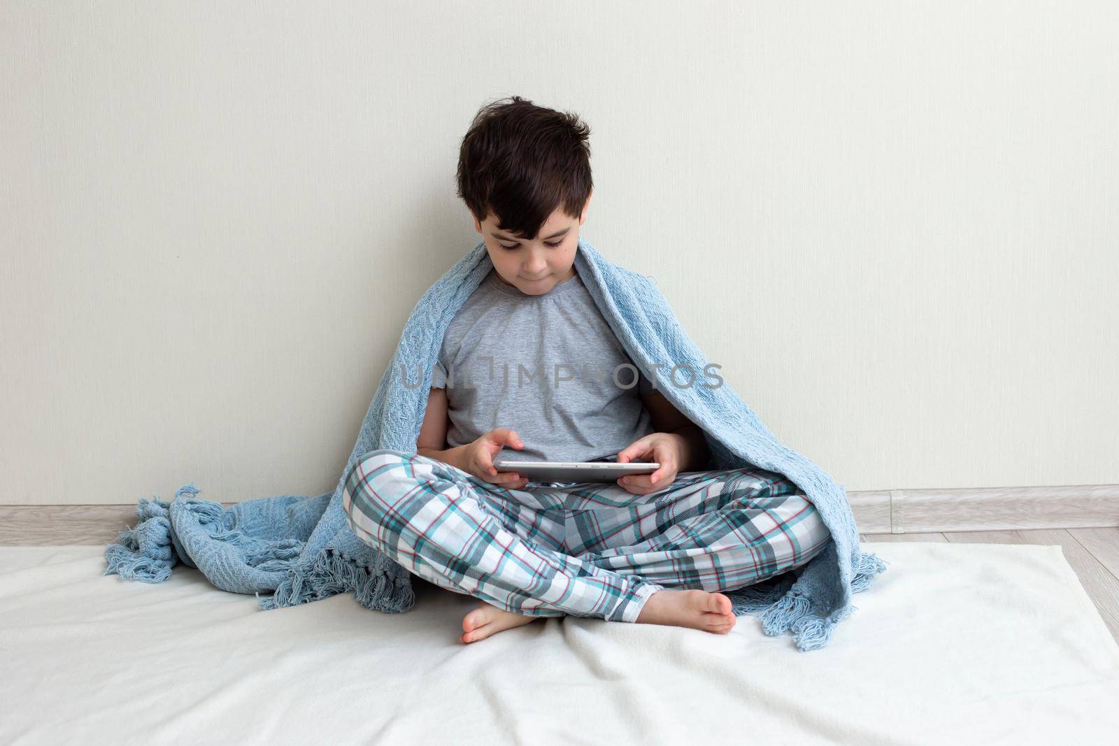 A brunette boy in gray pajamas, in a blue knitted plaid sits against a light wall, on the floor, uses a digital tablet. Copy space