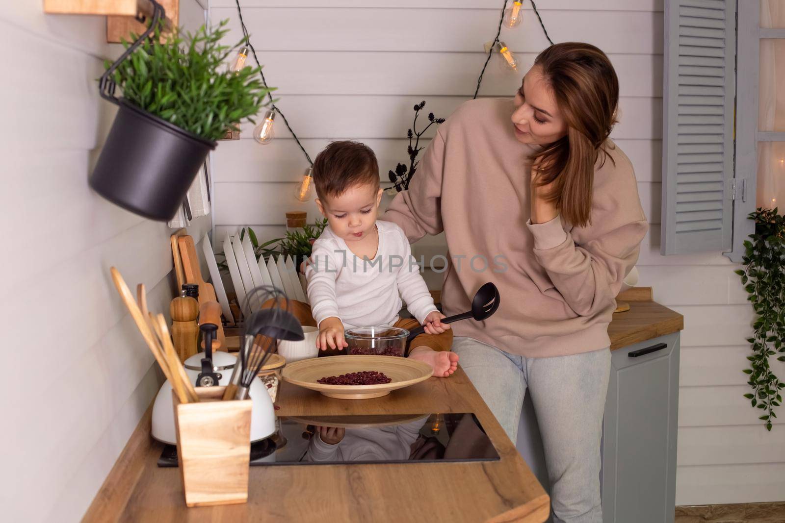 Young woman and little son play in the kitchen with dry red beans, together by Zakharova