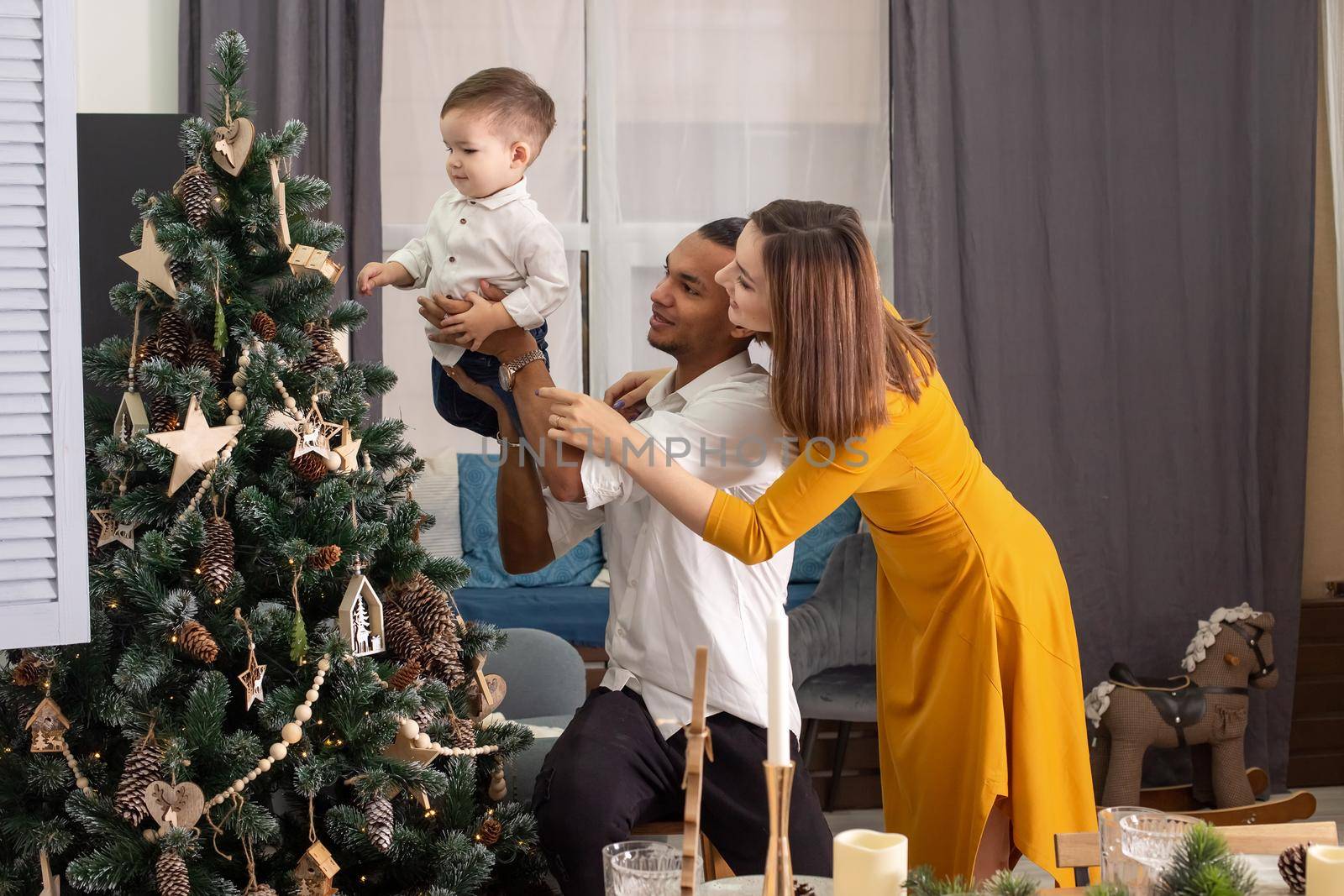 Happy young family: mulatto dad, little son and slender mom in mustard dress, show son a Christmas tree with wooden toys and cones, decor in eco style