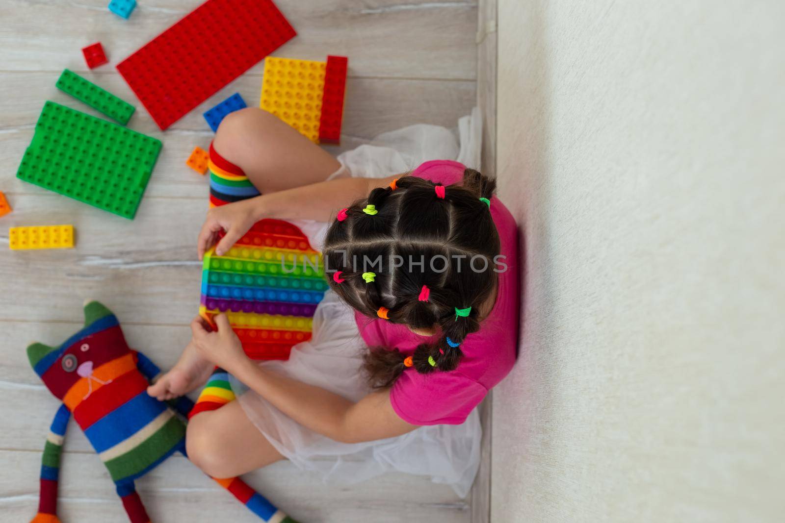 little brunette girl in bright colorful clothes, sits at home, on the floor, surrounded by colorful toys and rainbow silicone poppit. View from above