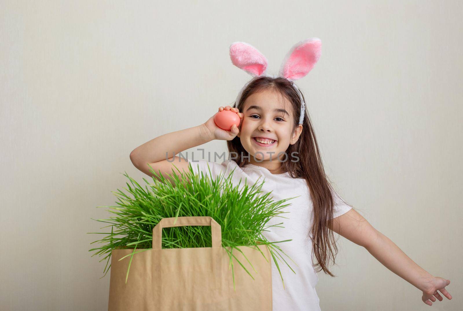 A little happy girl with hare ears, holds a pink egg and rejoices. Nearby is a paper bag with green grass. Close up. Copy space.