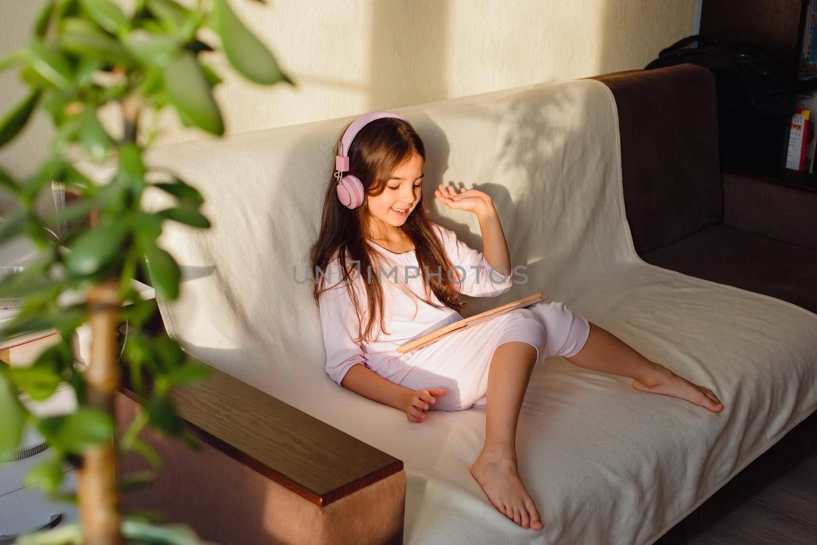 A friendly little beautiful girl in pink home clothes and pink headphones sits on the couch, communicates online using a digital tablet.