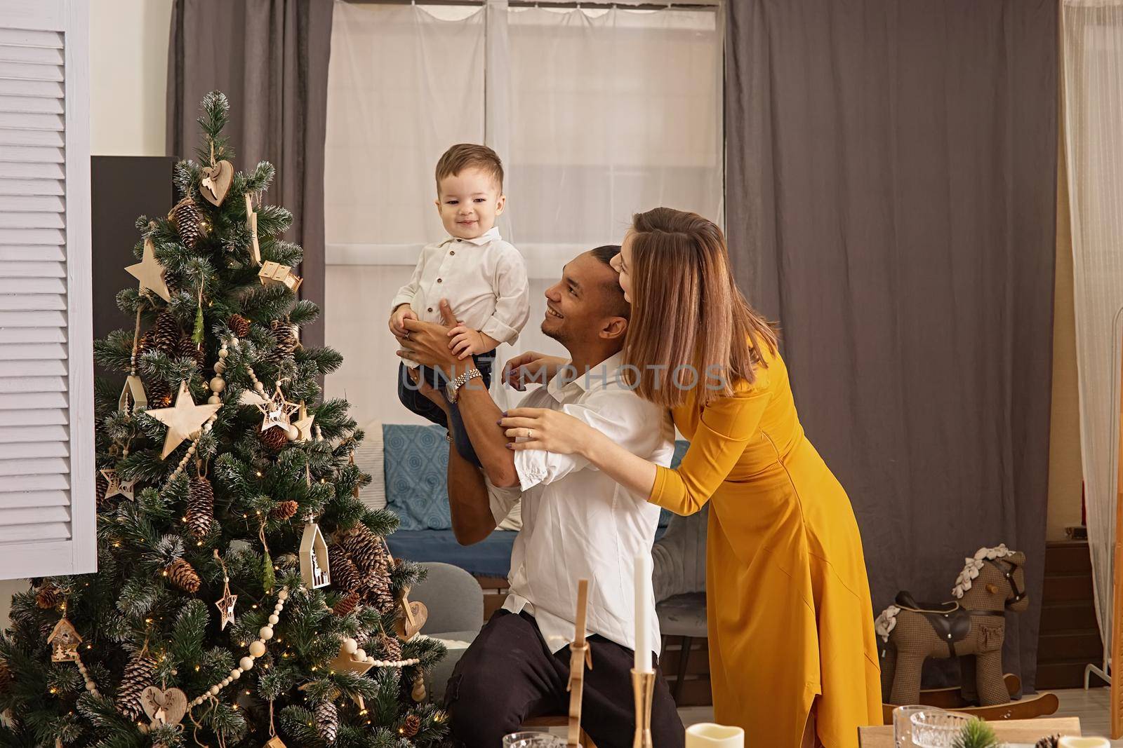 Happy young multiethnic family: dad, son and slender mom in mustard dress, show son a Christmas tree with wooden toys and pine cones, eco-style decor