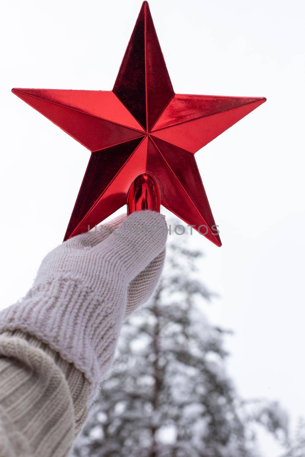 A hand in a white knitted mitten holds a red star over a snow pine tree in the forest. Vertical