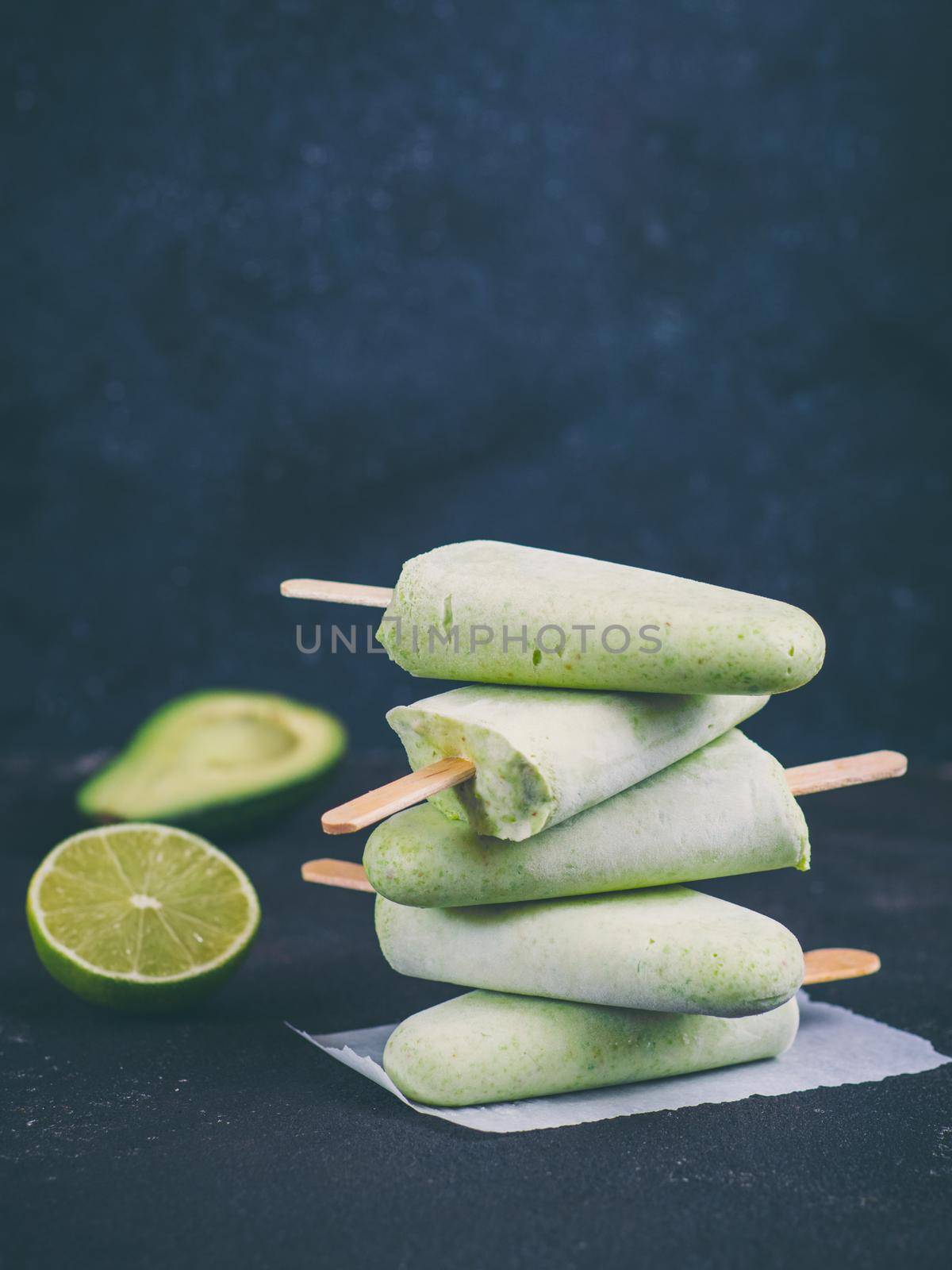 Homemade raw vegan avocado lime popsicle. Sugar-free, non-dairy green ice cream on dark blue textured background. Copy space. Ideas and recipes for healthy snack, dessert or smoothie. Vertical.