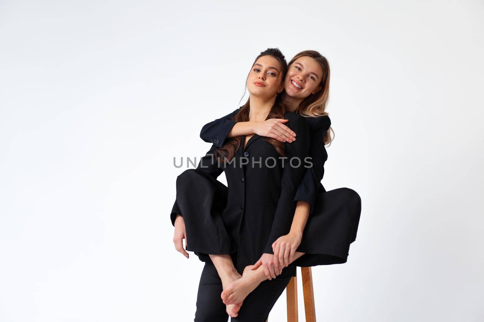 Two young business woman dressed black suit laugh and happy. One woman sitting chair and hugging other woman with barefoot legs by andreonegin