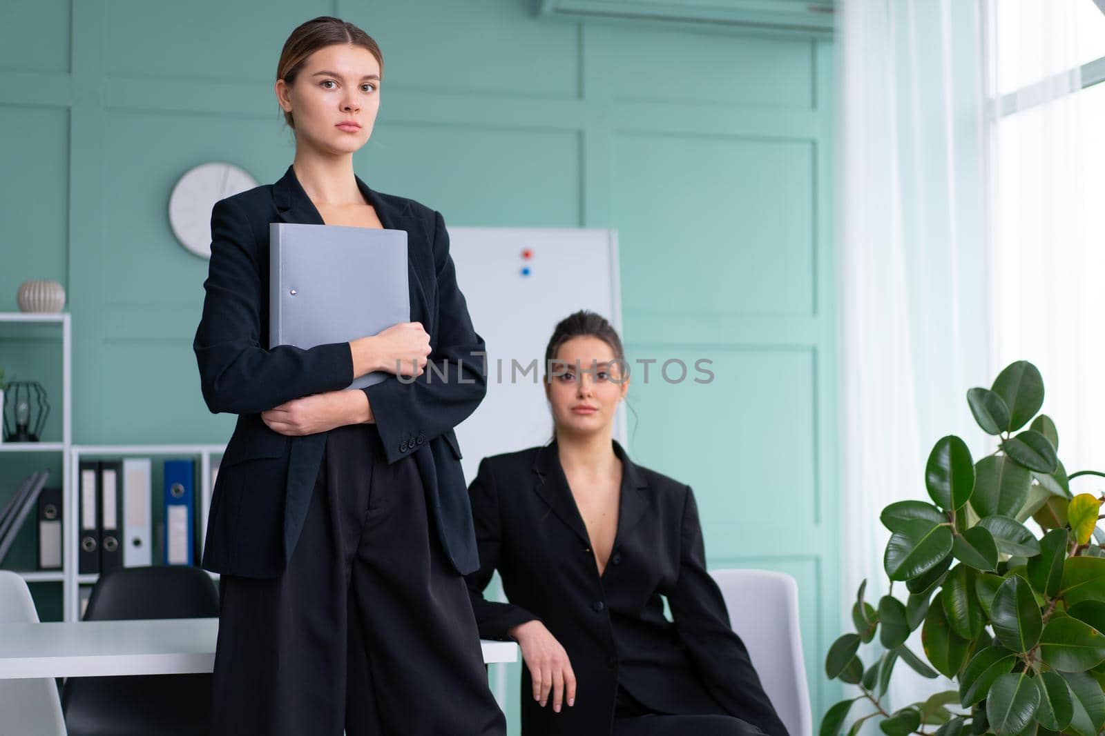 Young women leaders dressed black suit in office one woman standing with document folder other woman sitting on the table looking at camera. Business meeting. Two confidence caucasian female person.