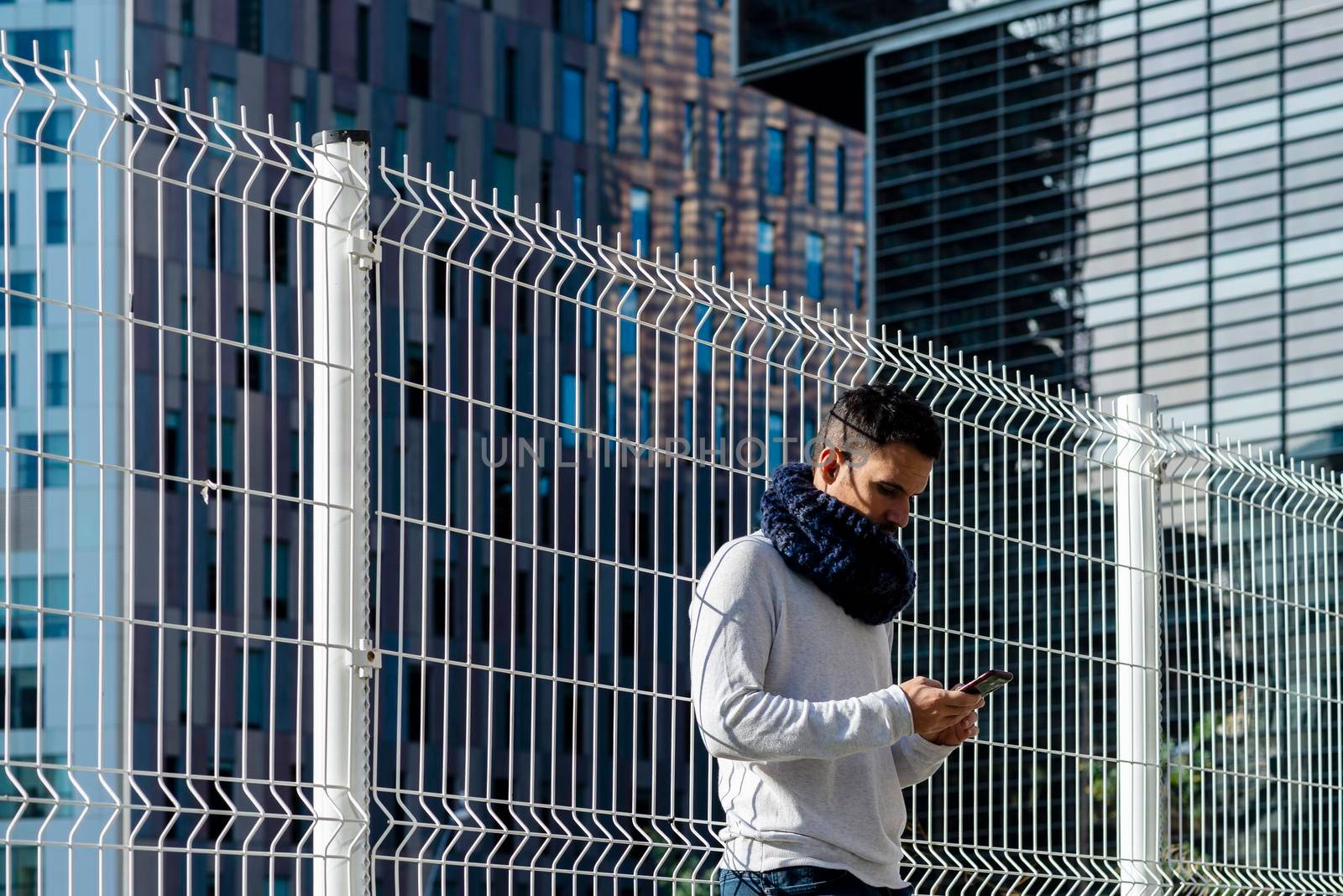 Young man wearing scarf leaning on metallic fence while using smartphone outdoors by raferto1973