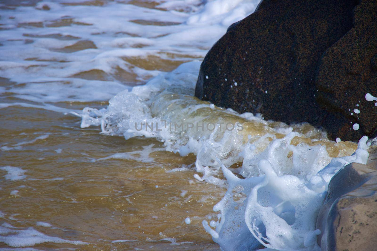 Macro photo of sea foam in motion as it crashes against dark rocks on sand beach with water droplets in the air. Norwich, Norfolk, taken with Canon EOS 90D