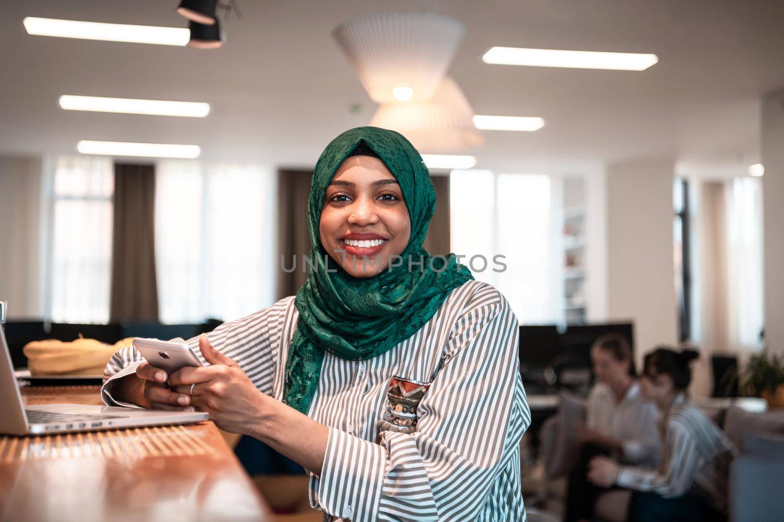 African Muslim businesswoman wearing a green hijab using smartphone while working on laptop computer in relaxation area at modern open plan startup office.High-quality photo
