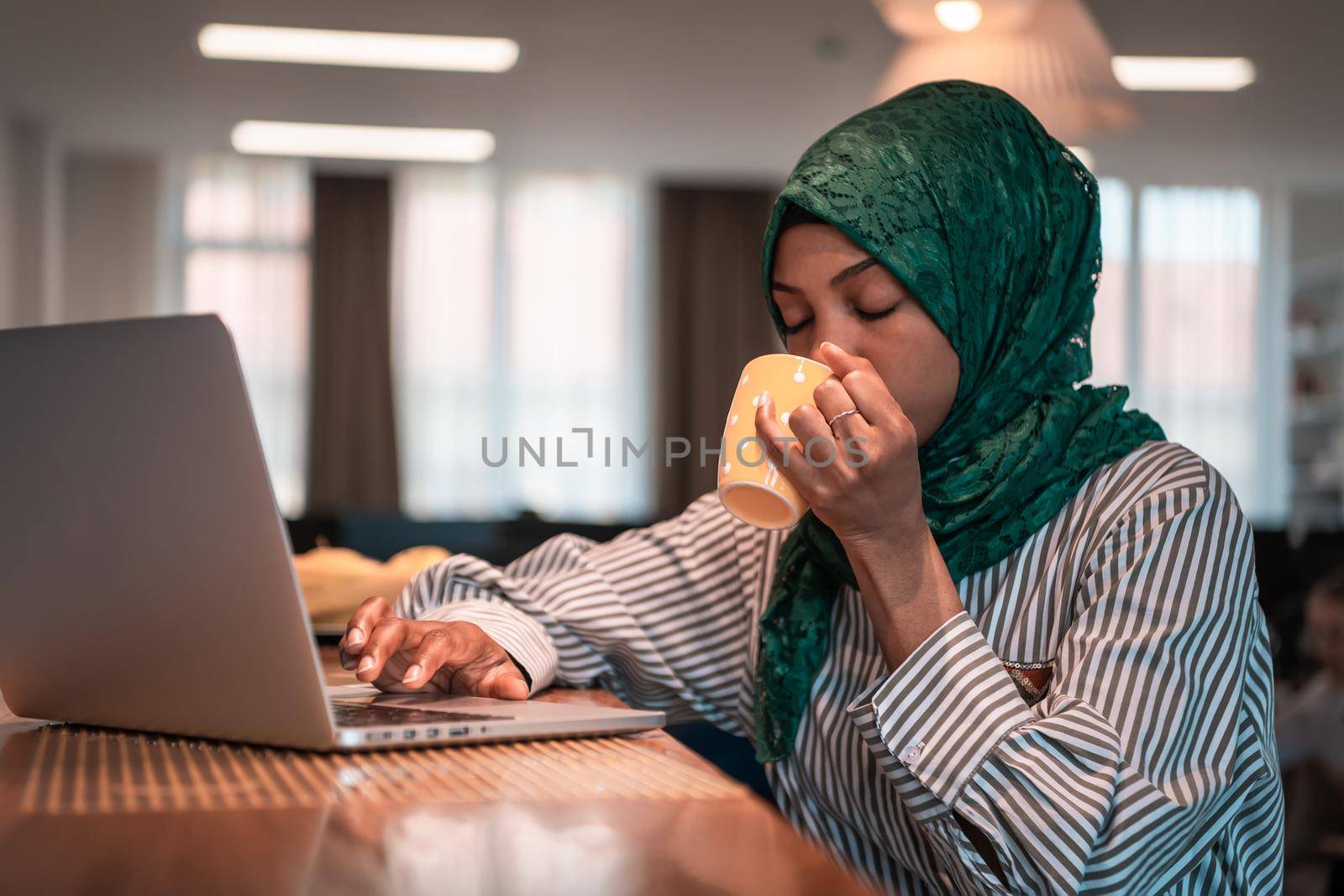 African muslim business woman wearing a green hijab drinking tea while working on laptop computer in relaxation area at modern open plan startup office. by dotshock
