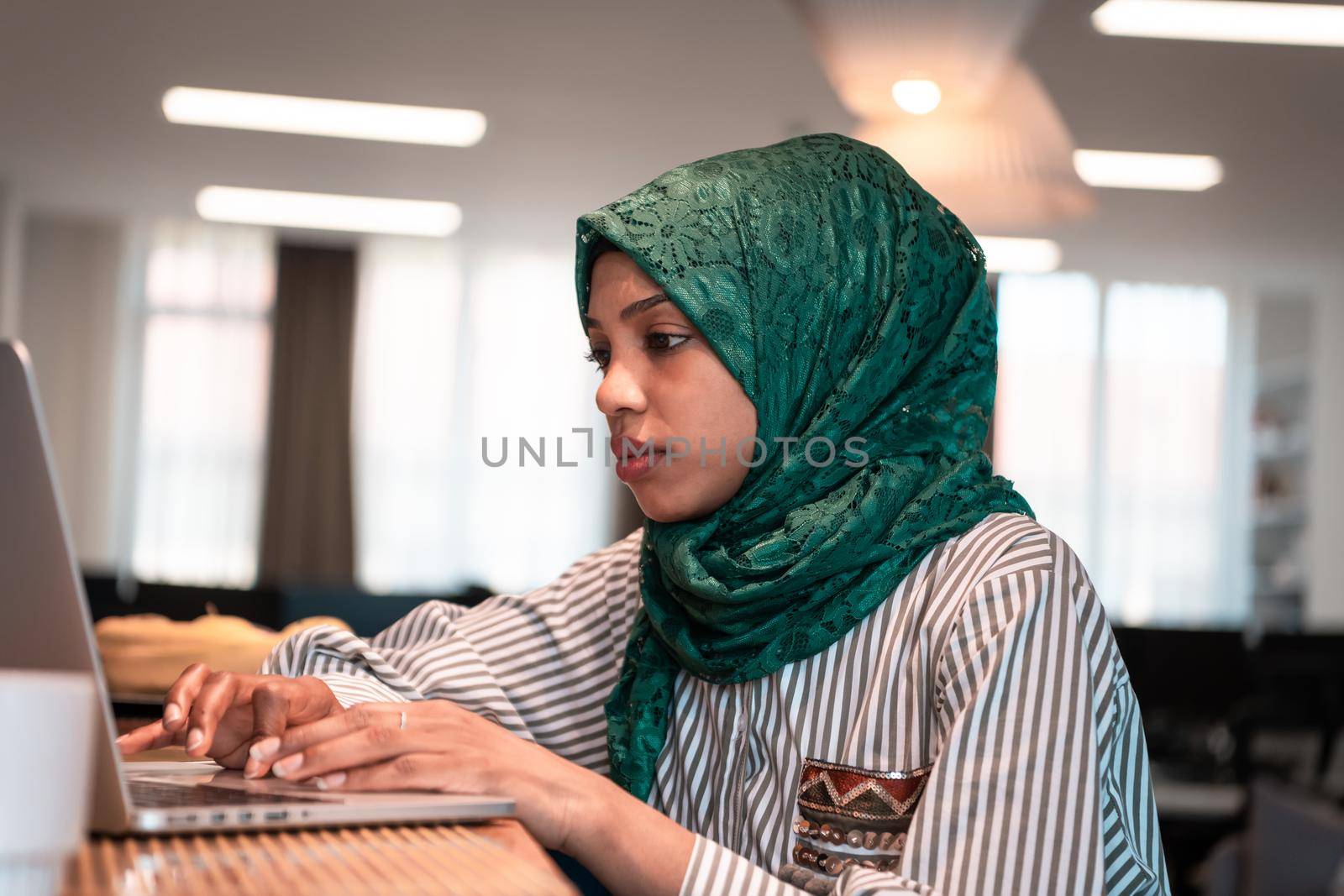 African Muslim businesswoman wearing a green hijab and working on laptop computer in relaxation area at modern open plan startup office. High-quality photo